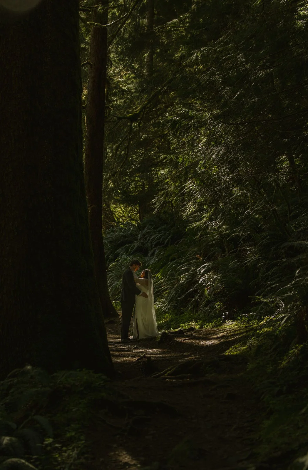 A couple dressed in wedding attire standing in a lush green forest, embracing each other on a dirt path surrounded by tall trees and dense foliage.