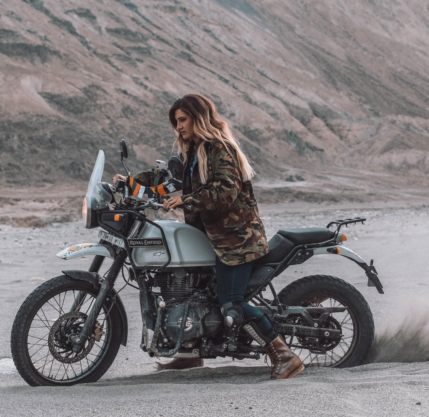 A woman in military-style camouflage jacket and brown boots standing next to a silver Royal Enfield motorcycle in a desert landscape with mountains in the background.