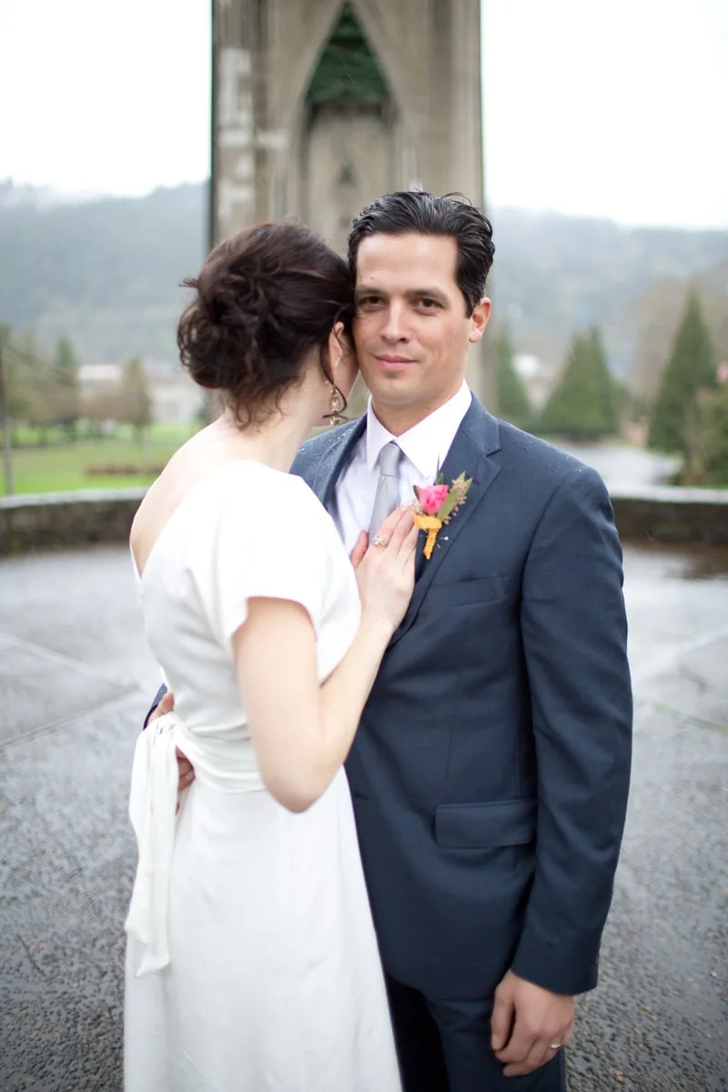A bride and groom standing close together outdoors, with a bridge and river in the background.
