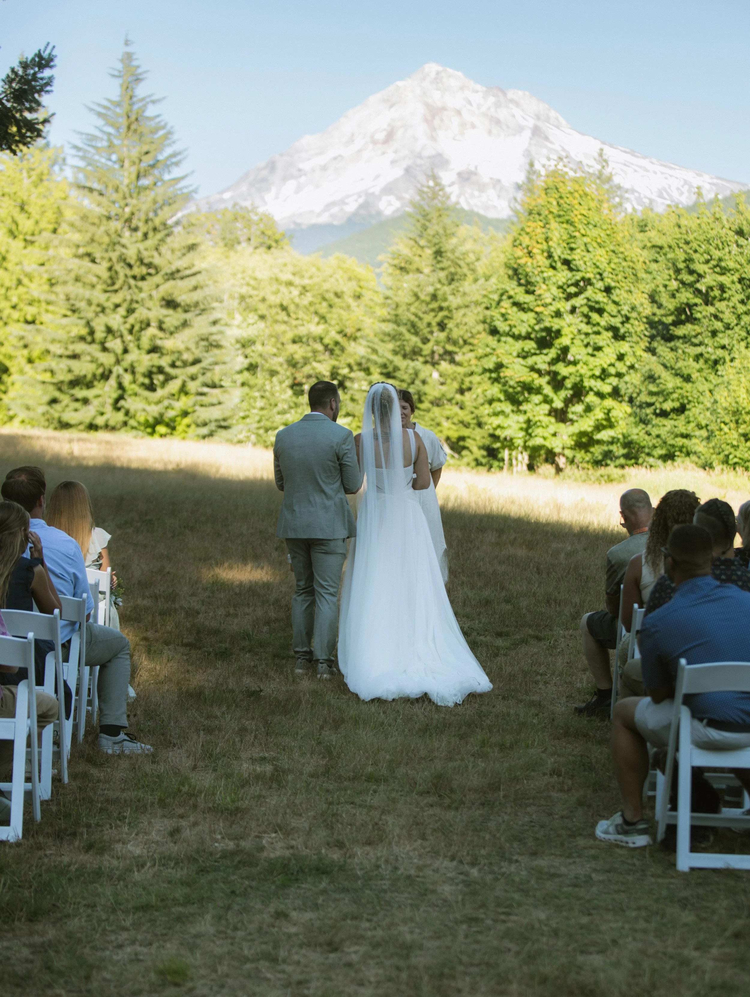 A wedding ceremony outdoors with a mountain and trees in the background. A bride and groom stand facing each other, surrounded by seated guests.