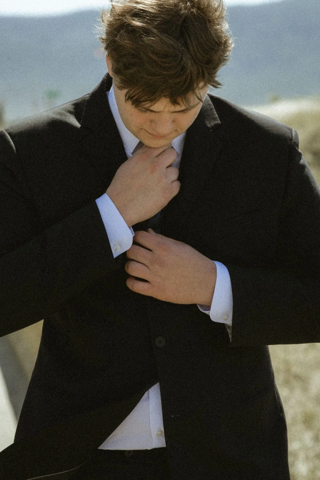 A young man with curly brown hair dressed in a black suit with a white shirt, looking down and holding his own collar with both hands outdoors near a body of water.