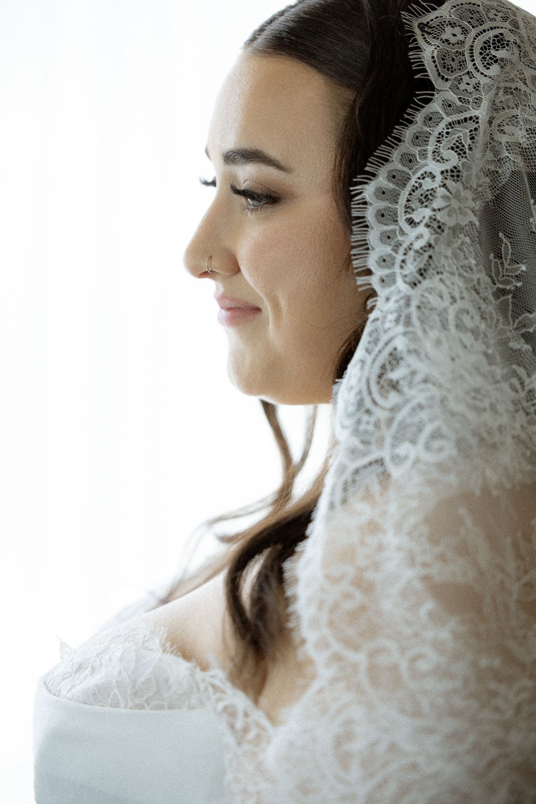 Side profile of a woman with dark hair, wearing a lace head covering and a lace dress, with a serene smile.