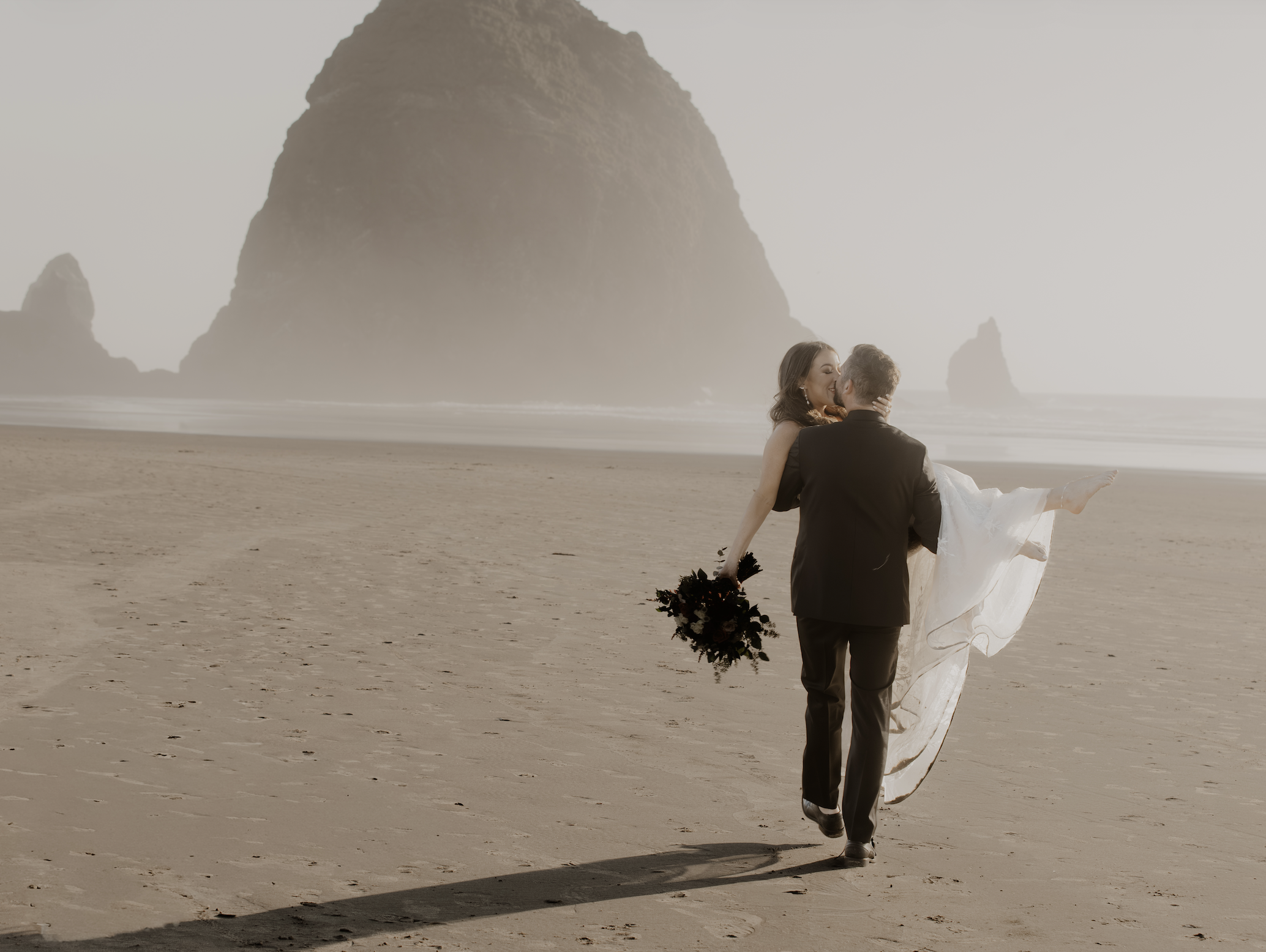 A couple in wedding attire walking on a beach with a large rock formation in the background. The bride is holding a bouquet and being carried by the groom.