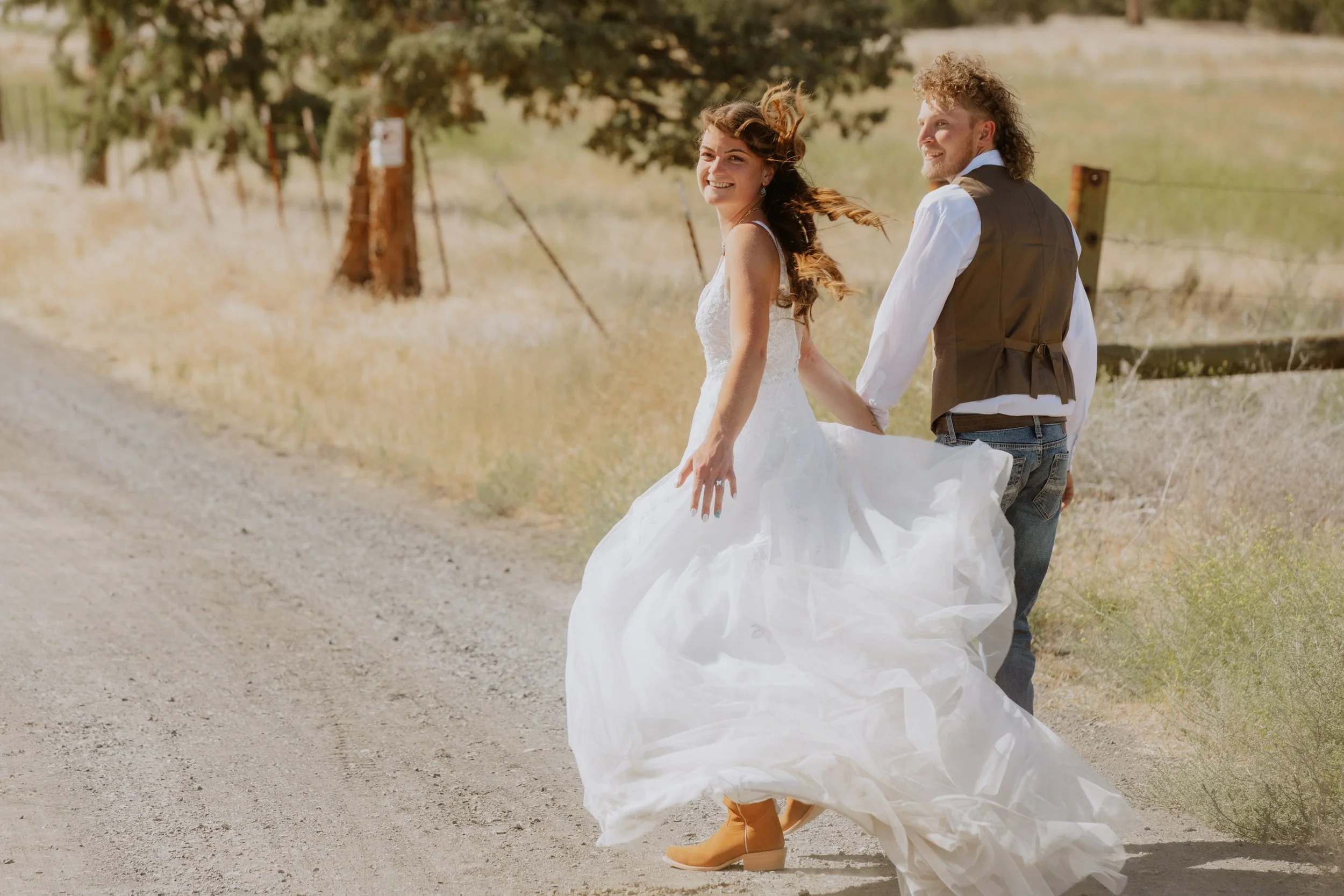 A bride and groom walking outdoors on a sunny day, with the bride wearing a white wedding dress and tan boots, and the groom wearing jeans, a white shirt, and a brown vest, holding hands and smiling.