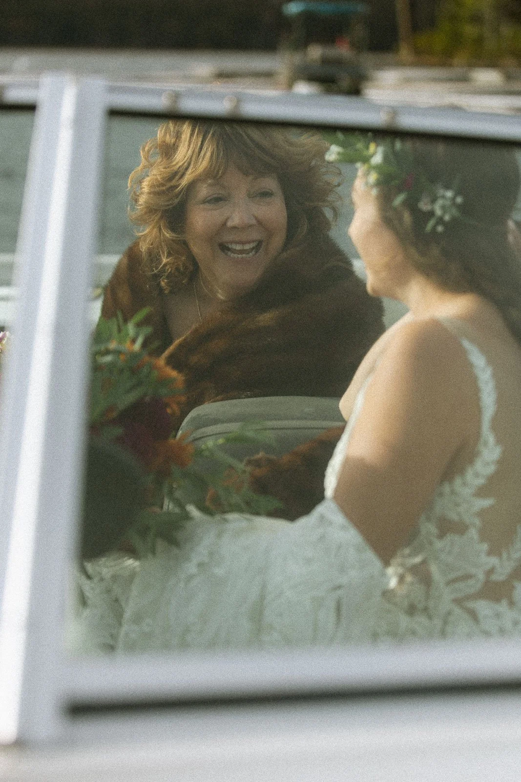 A woman in a wedding dress with a floral headpiece is riding in a vehicle with another woman, who is smiling at her.
