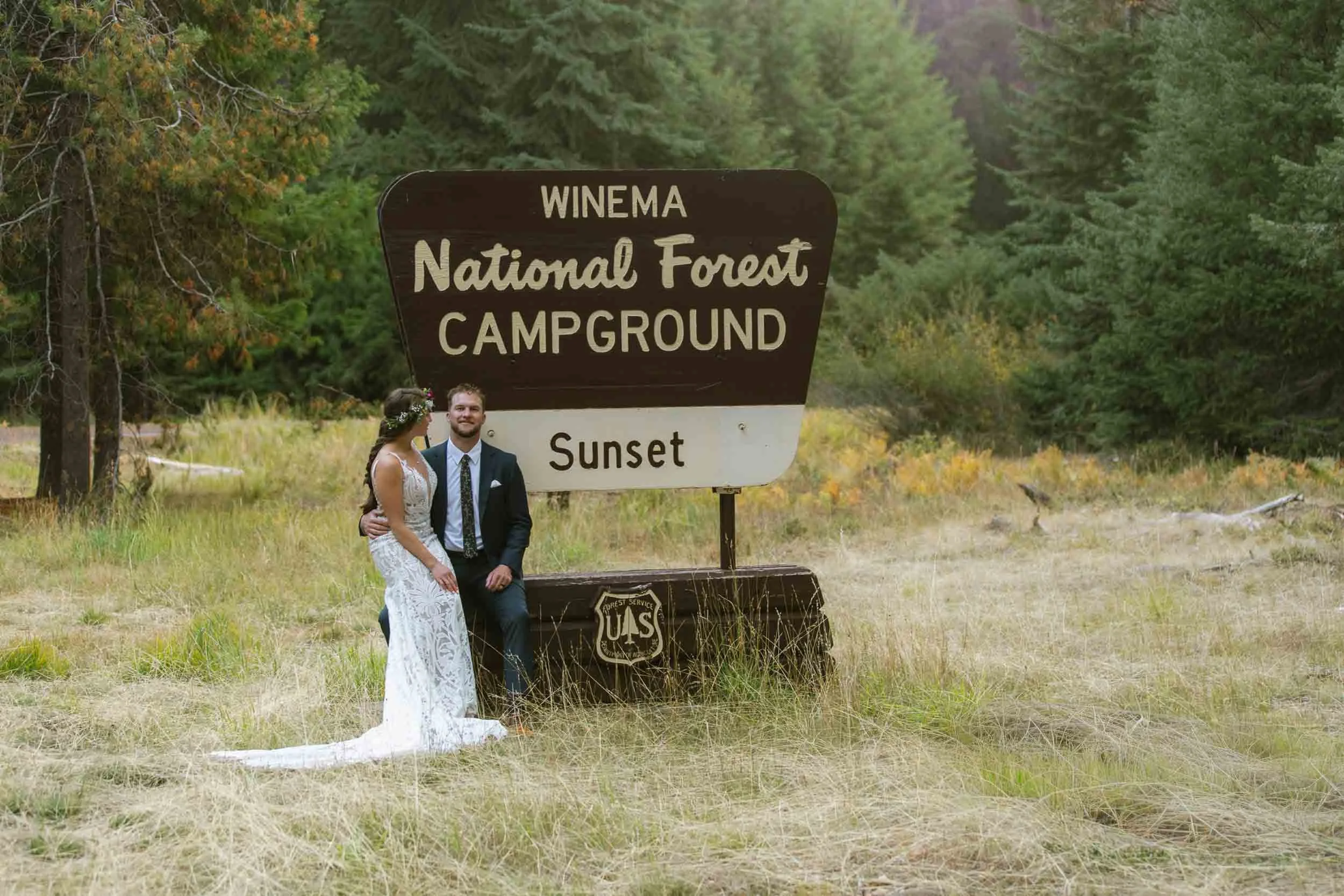 A newlywed couple in wedding attire, standing and sitting next to a large sign that reads "Winema National Forest Campground Sunset," with trees and grass in the background.