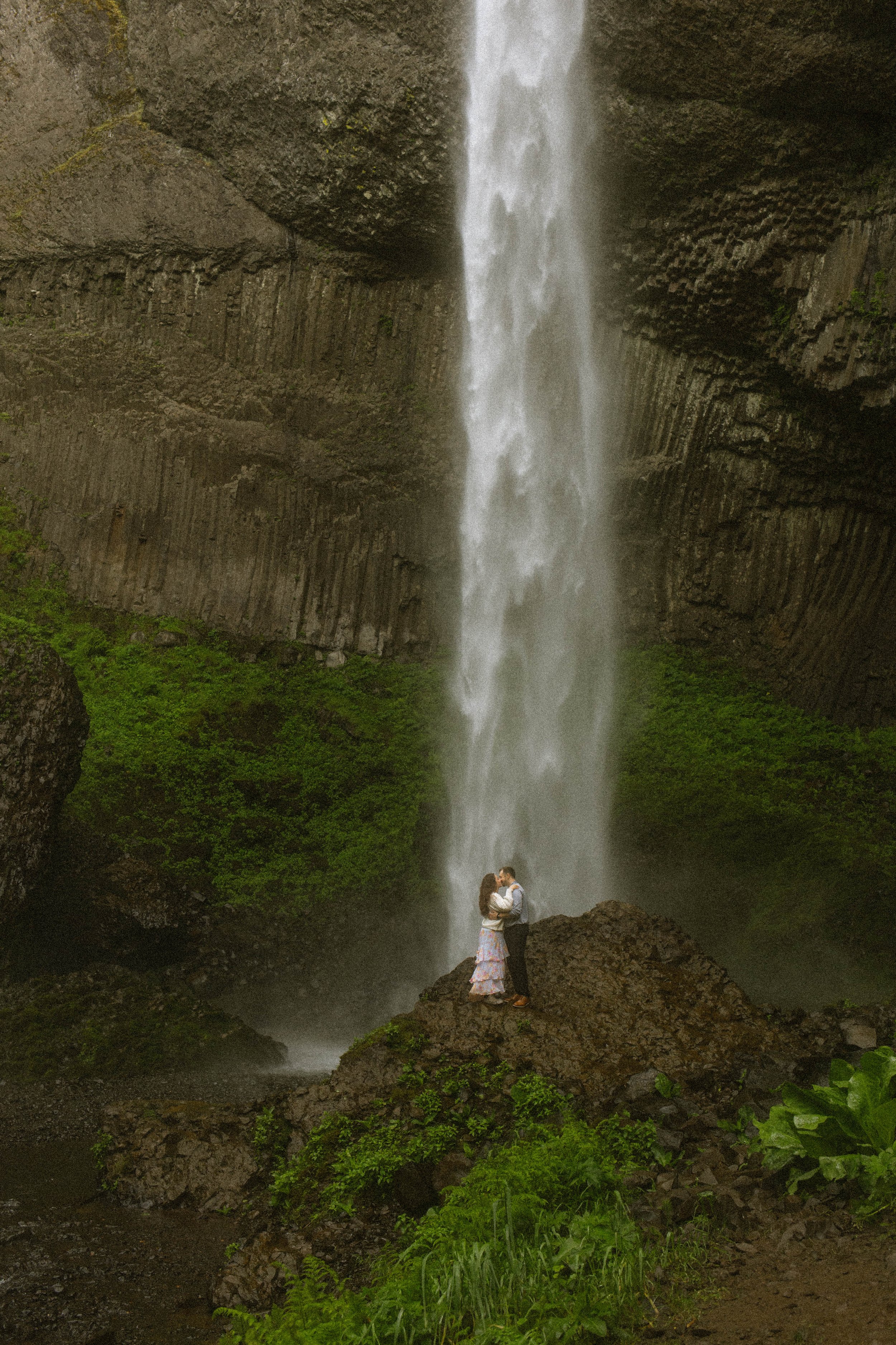 A couple standing on a large rock in front of a tall waterfall with lush green moss and foliage around.