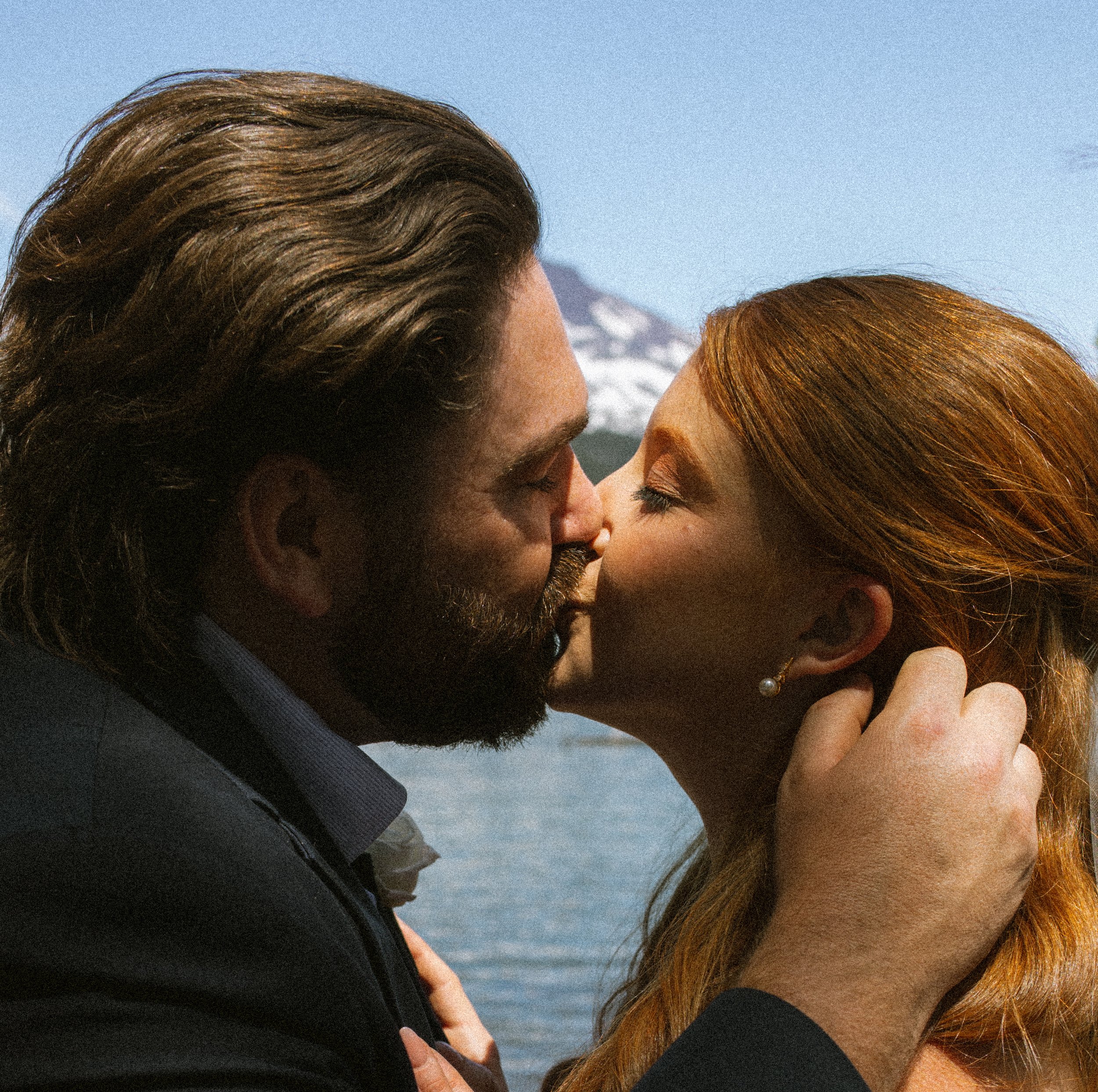 A man and woman are sharing a kiss outdoors in front of a snow-capped mountain and water.