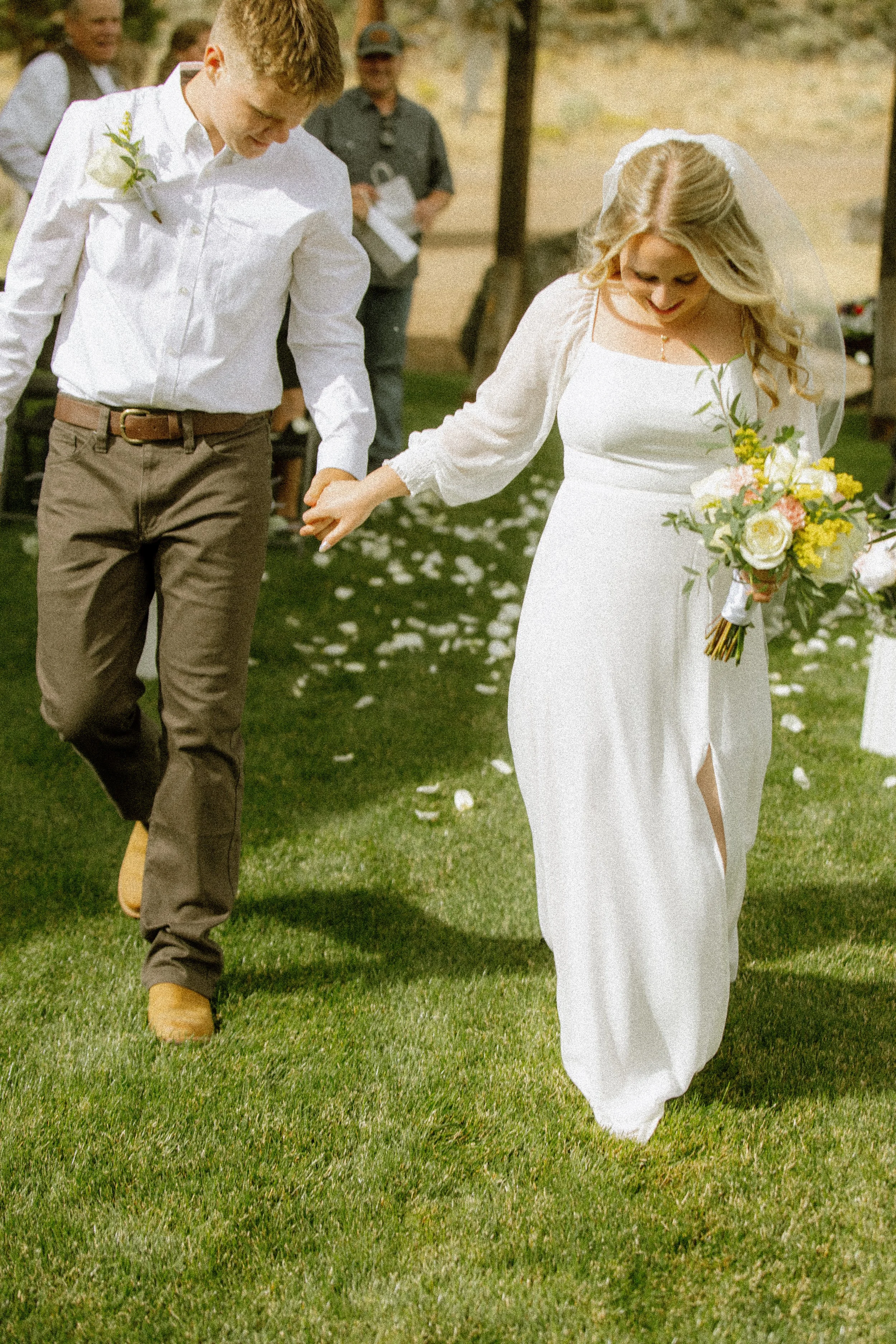 A bride and groom holding hands and walking on grass during their wedding celebration, with guests in the background.