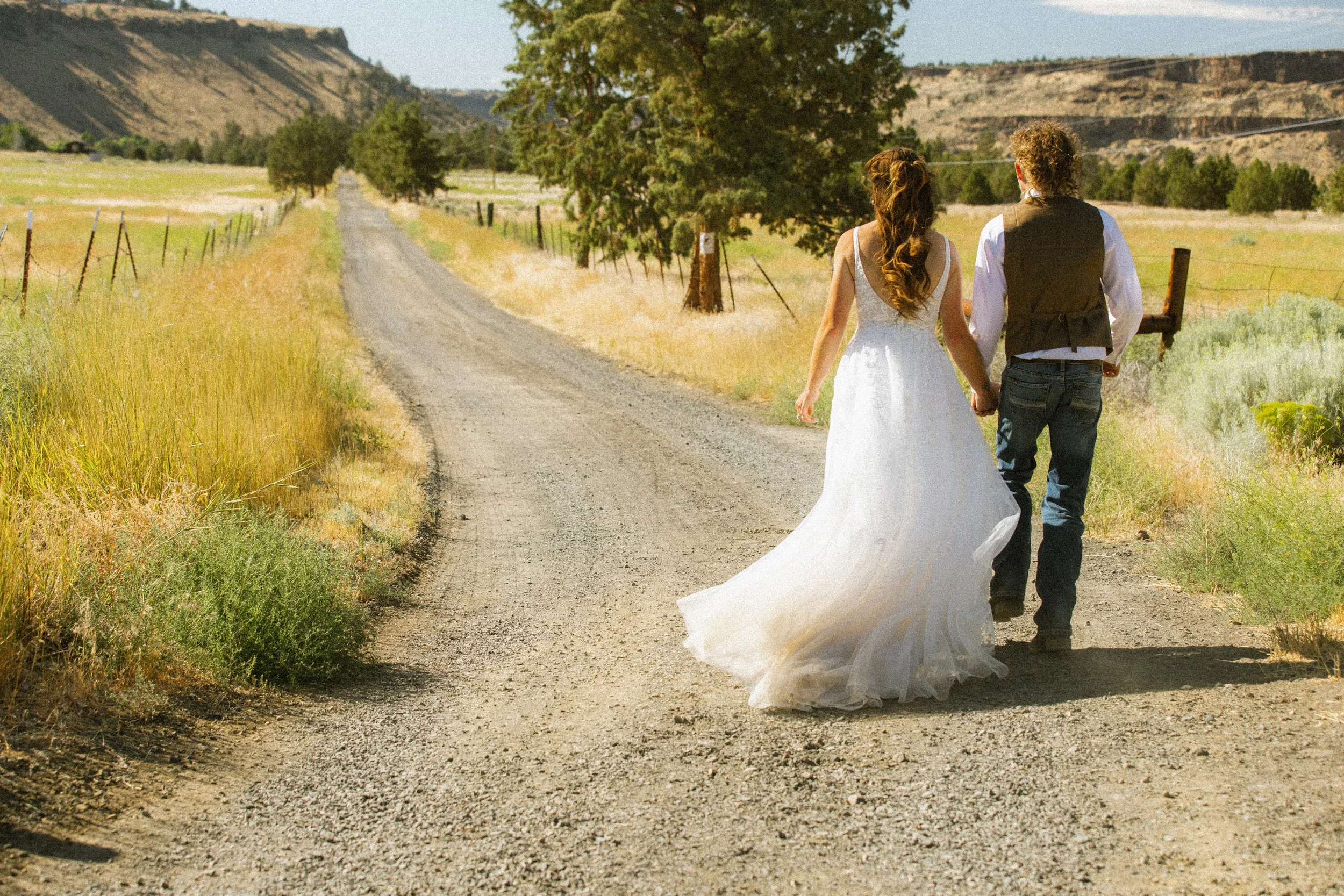 A bride and groom walking hand in hand on a dirt road in a rural landscape, with mountains and trees in the background.