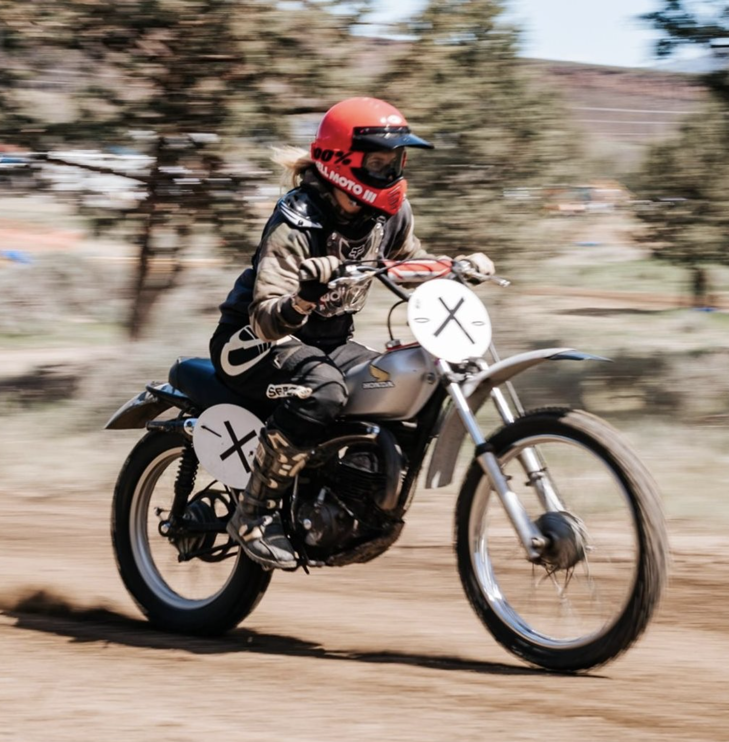 Person riding a dirt bike on a dirt track, wearing a red helmet and protective gear, with a blurred background indicating motion.