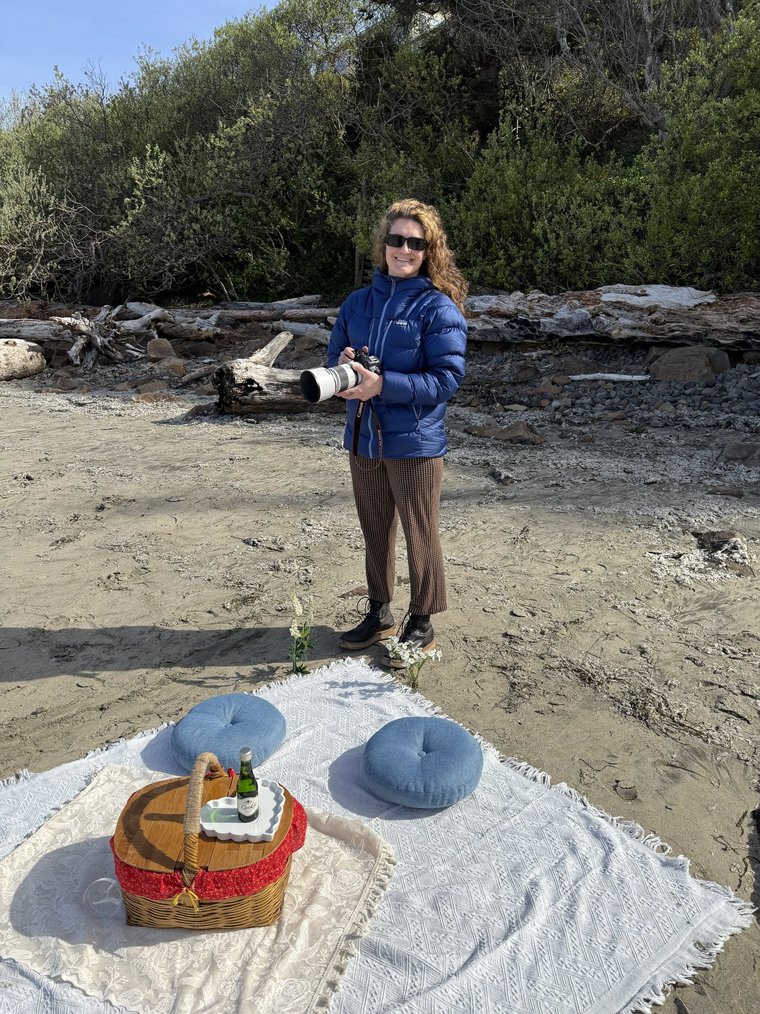Woman standing on a sandy beach holding a camera, with a picnic setup on a white blanket in front, featuring a basket, two blue cushions, a tray with drinks, and small flowers, with trees in the background.