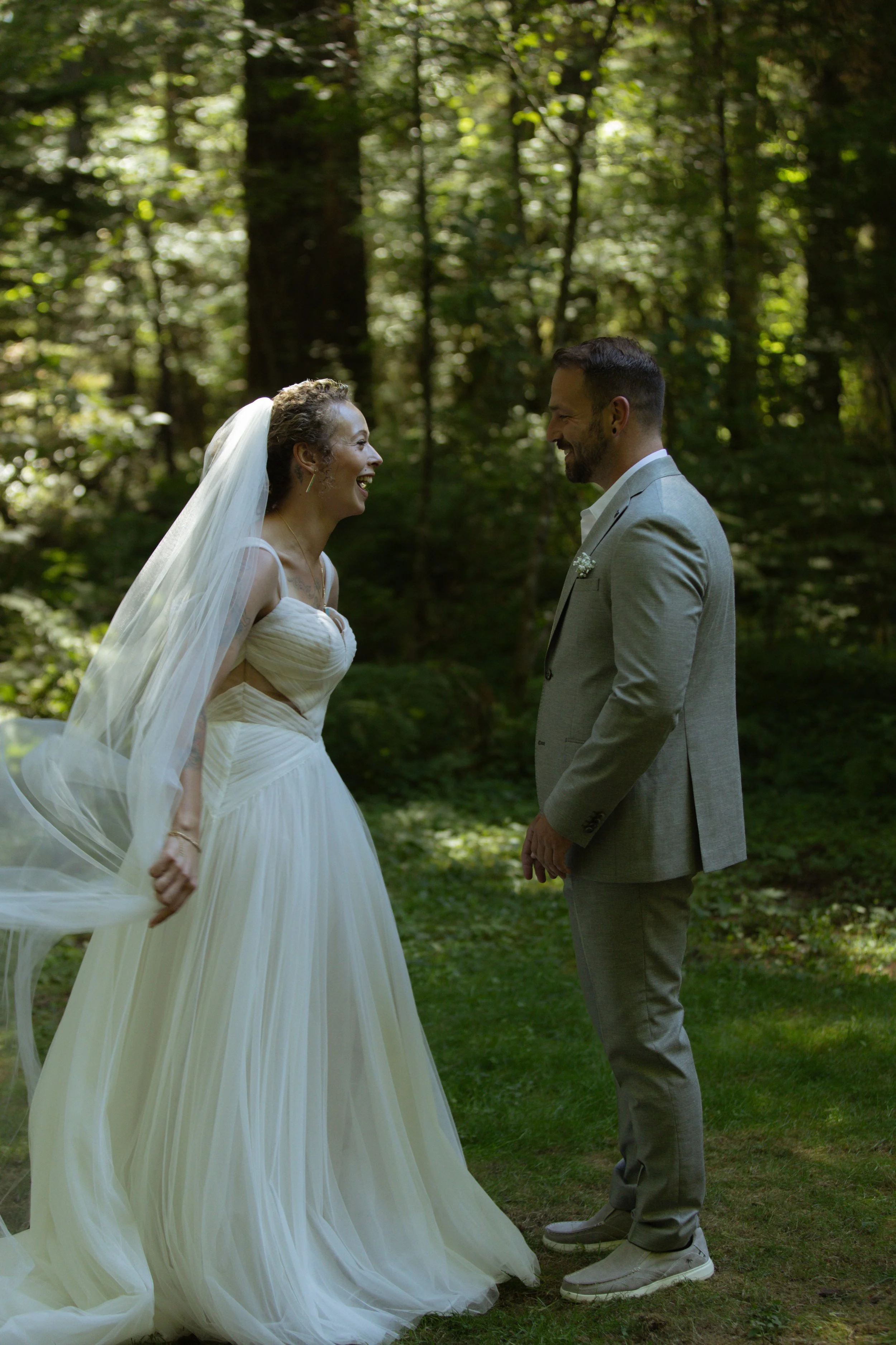 A bride and groom smiling at each other in a forest, with the bride wearing a white wedding dress and veil, and the groom in a light gray suit.