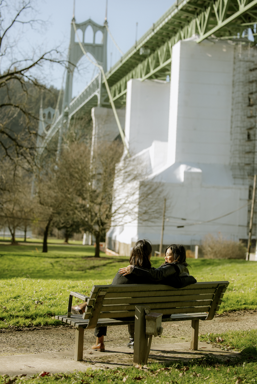 Two women sitting on a park bench enjoying a sunny day, with the suspension bridge in the background.