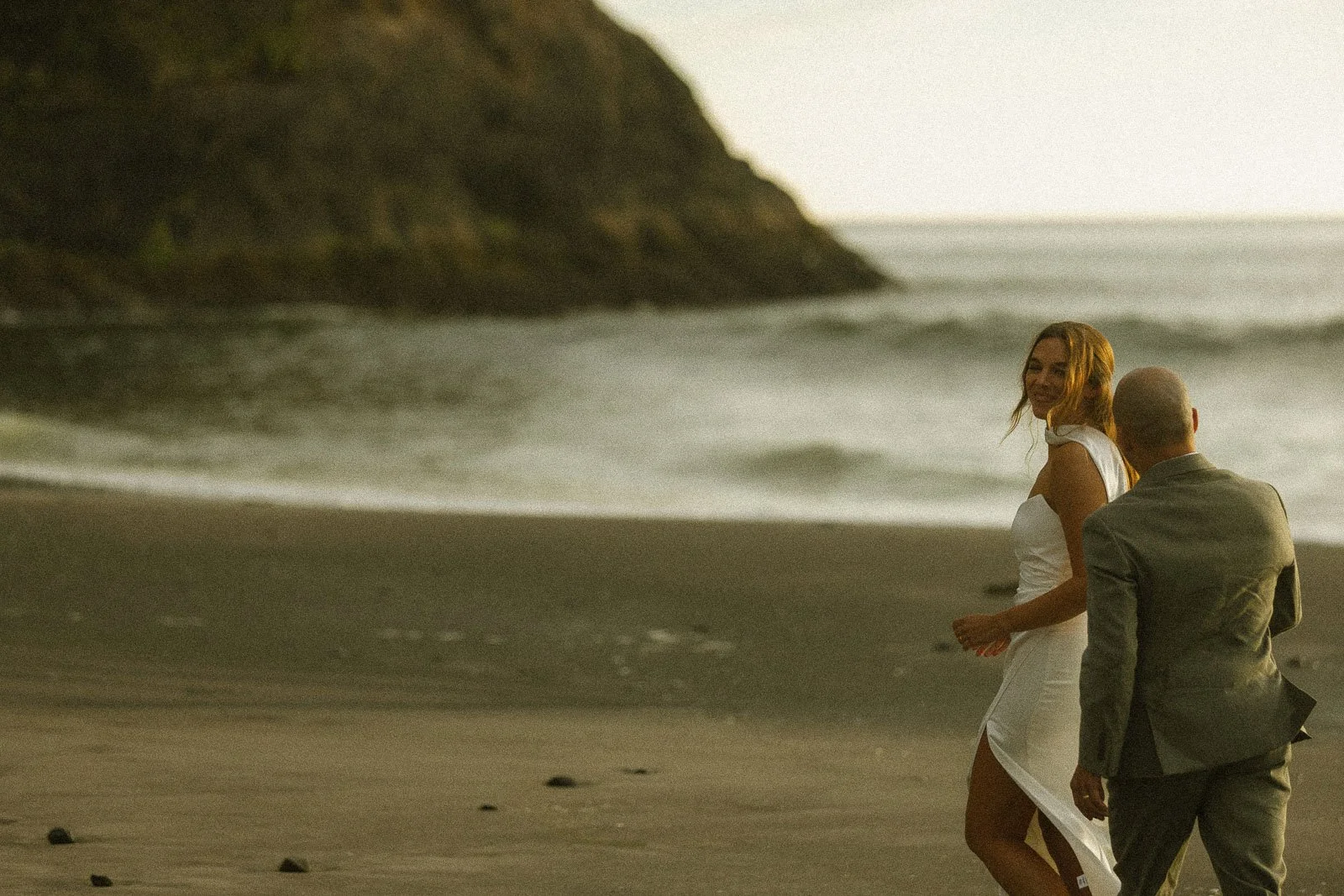 A couple in wedding attire walking along the beach with waves in the background, smiling and enjoying moment together.