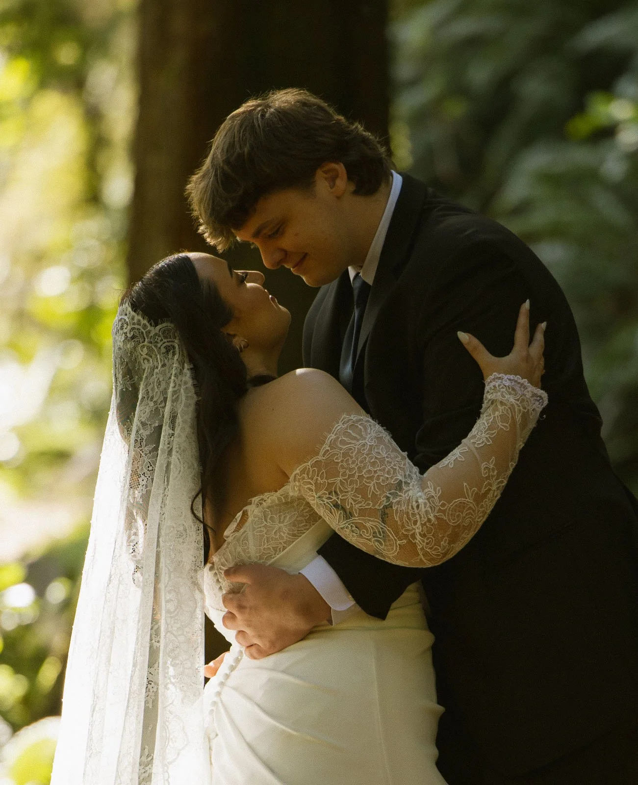 A bride and groom share a kiss outdoors in a wooded setting, with the bride wearing a lace wedding dress and veil, and the groom in a black suit. They are embracing, smiling, and looking into each other's eyes.