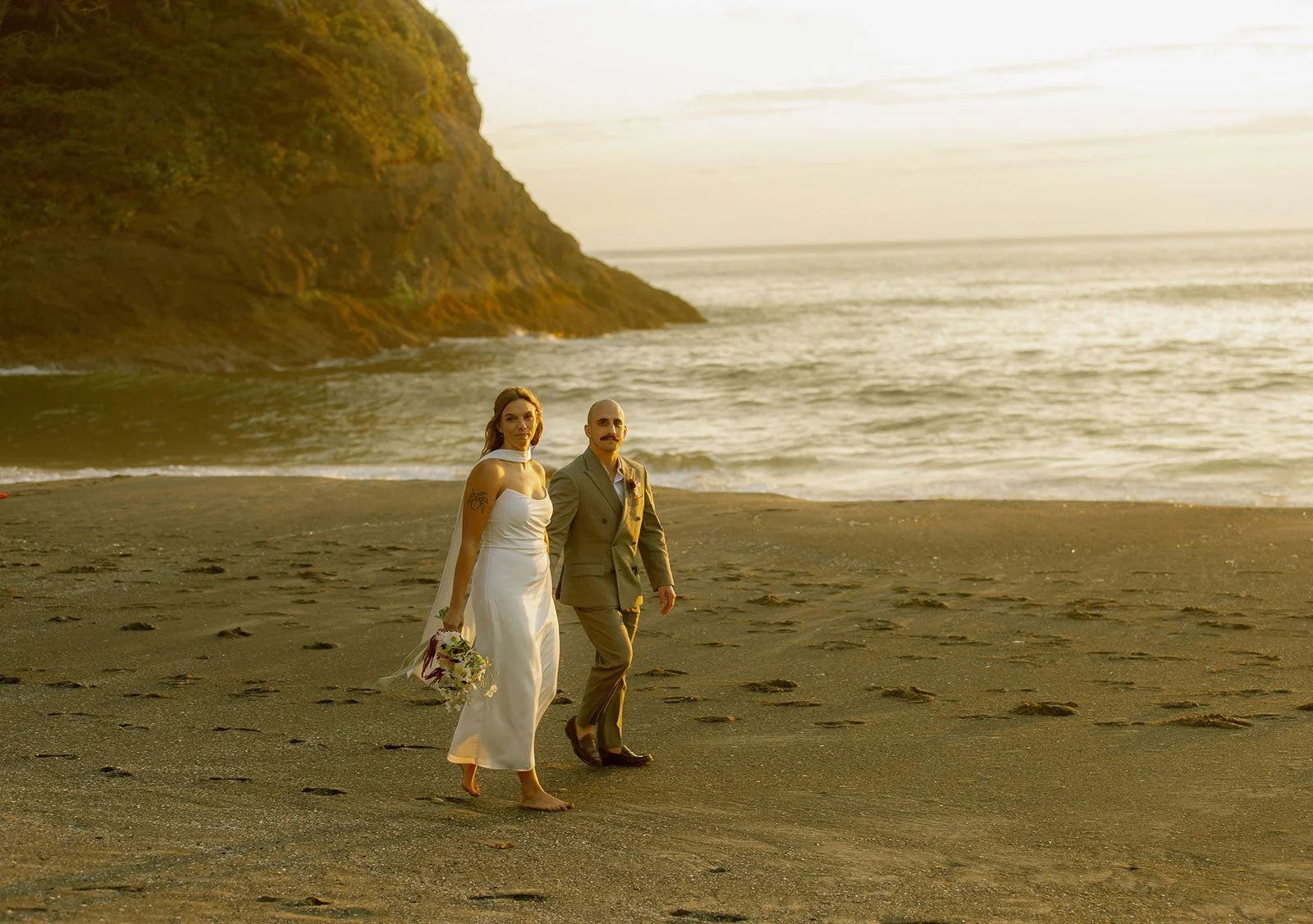 A couple walking on a sandy beach during sunset, with a woman in a white dress holding a bouquet and a man in a suit, near a large cliff by the ocean.