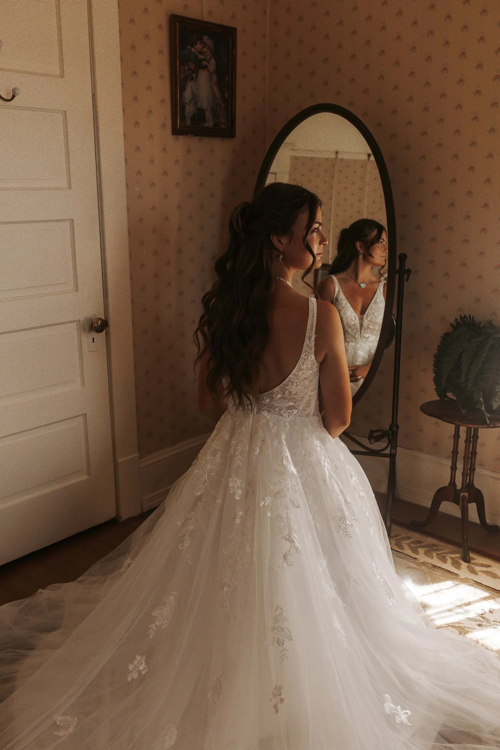 A woman in a white wedding gown standing in front of a mirror, looking at her reflection.