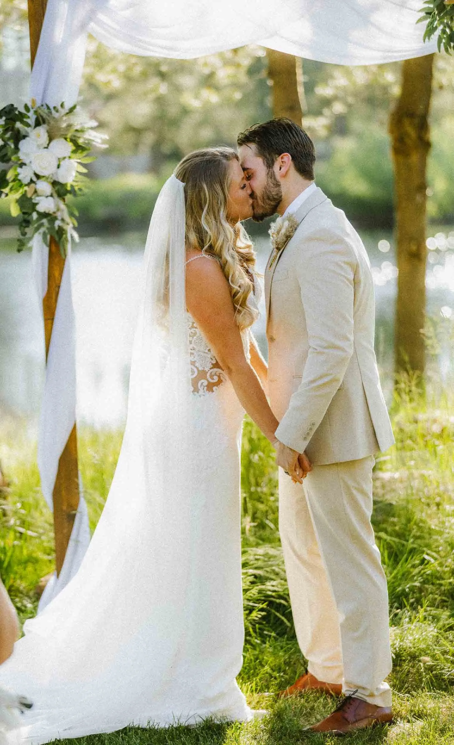 A bride and groom stand in a grassy outdoor setting with water and trees in the background, illuminated by sunlight, during their wedding ceremony.