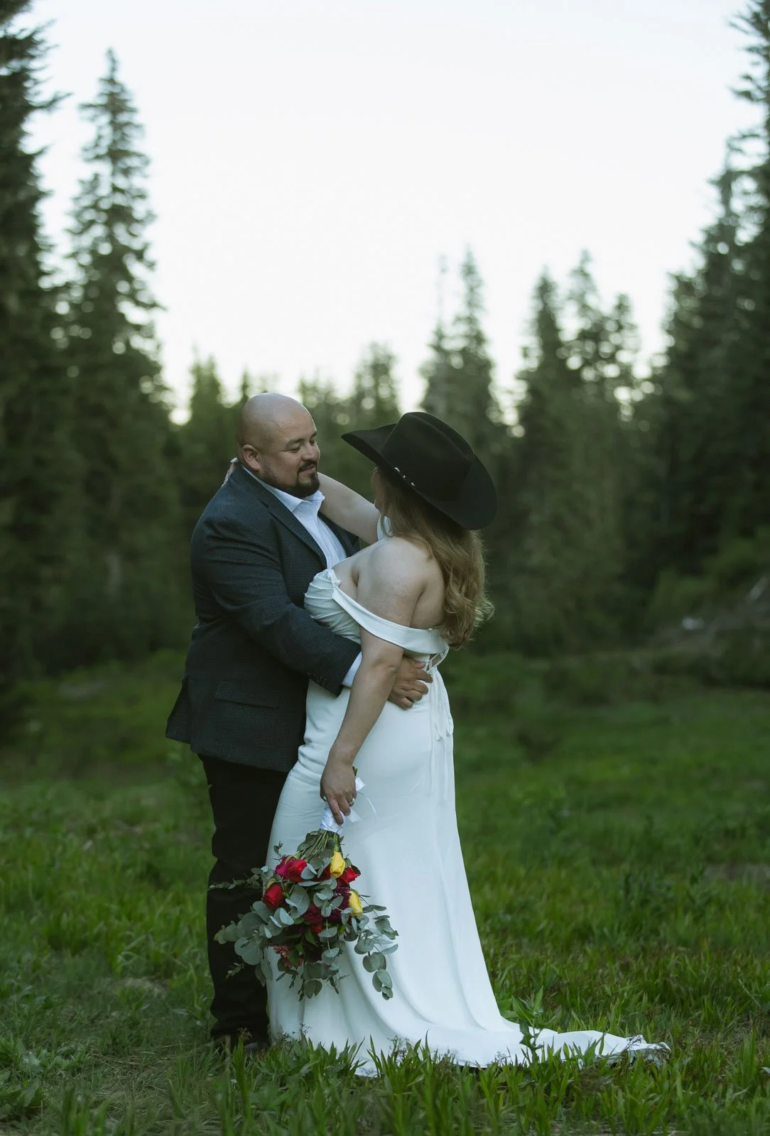 A couple dressed in wedding attire standing outdoors in a grassy field with pine trees in the background, the man wearing a dark suit and the woman in a white off-shoulder dress with a black wide-brimmed hat, holding a bouquet of red, yellow, and gray flowers.