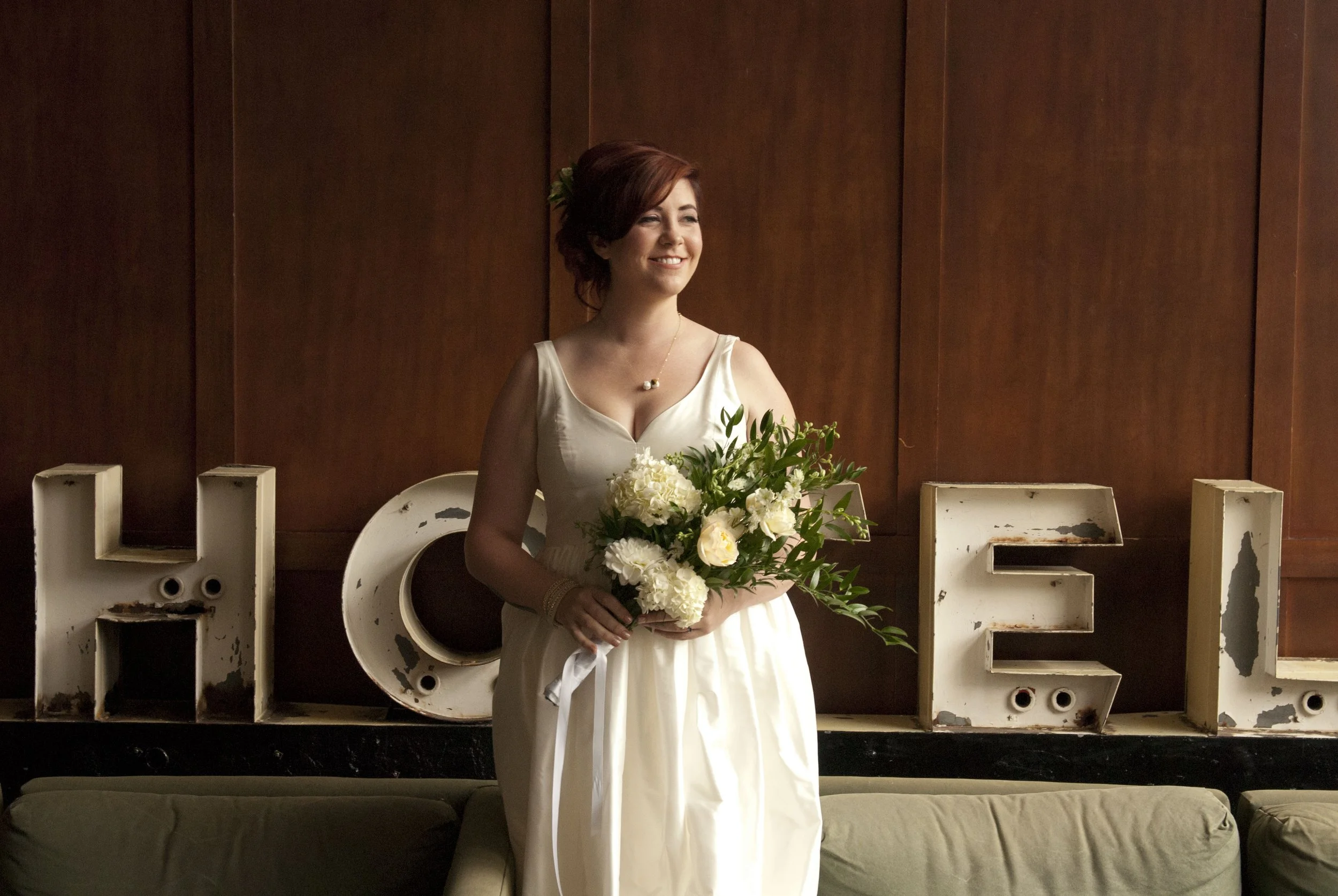 A woman in a white dress holding a bouquet of white and green flowers, smiling indoors near vintage letters spelling 'HOME' on a wall.