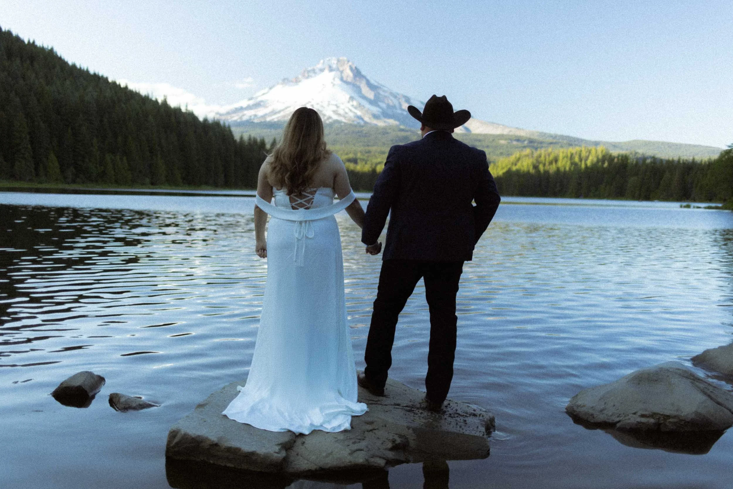 A couple holding hands standing on rocks in a lake, with a snow-capped mountain in the background.