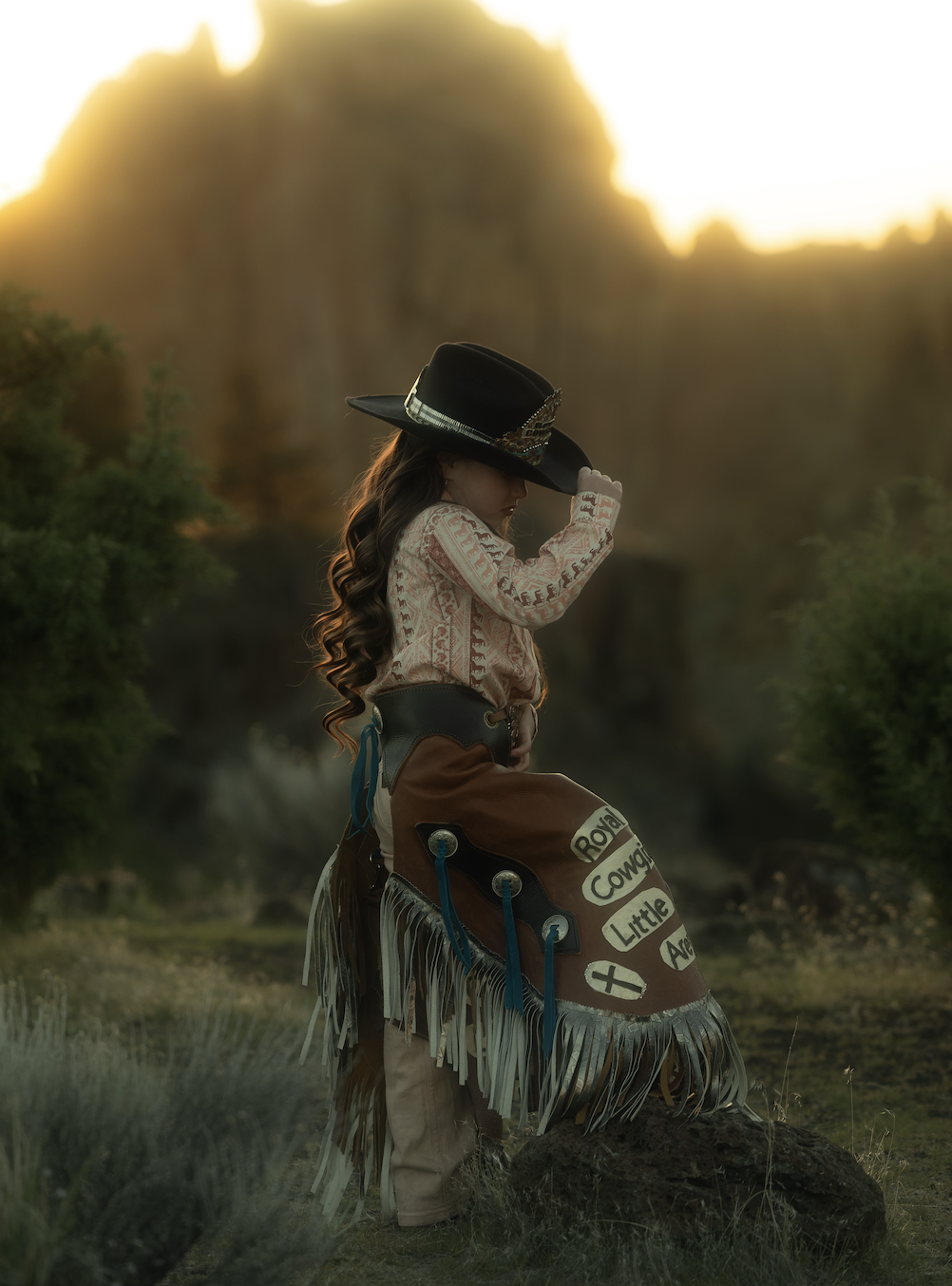 A young girl dressed as a cowboy, wearing a wide-brimmed hat, long-sleeved shirt, and fringed vest, standing outdoors at sunrise or sunset. She is standing on a rock with her head bowed and hat held shading her face, in a natural landscape with green
