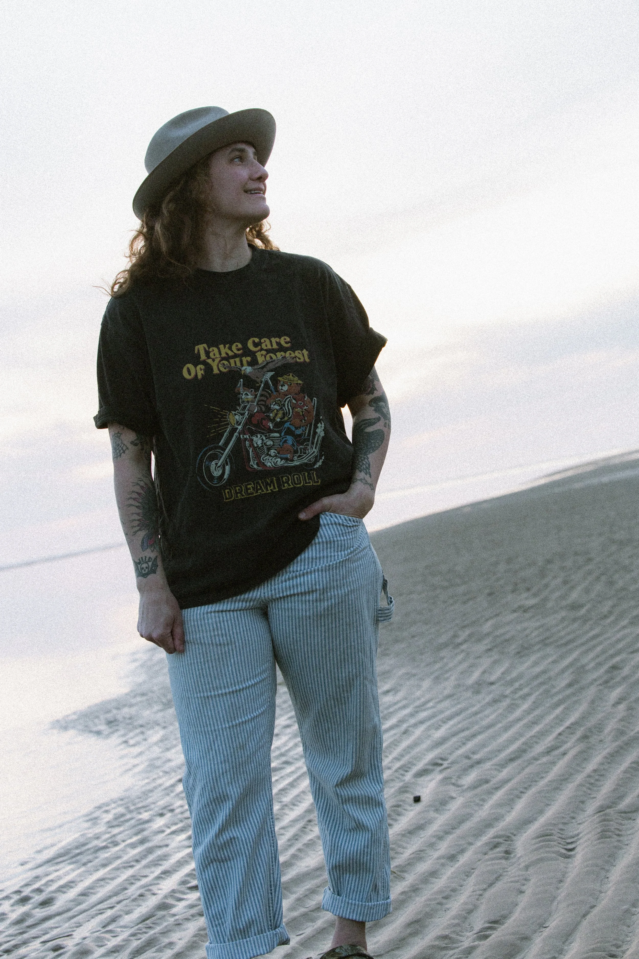 Woman standing on beach, wearing a black T-shirt with graphic print, light blue striped pants, a grey wide-brimmed hat, and has tattoos on her arms, looking at the sky at sunset.