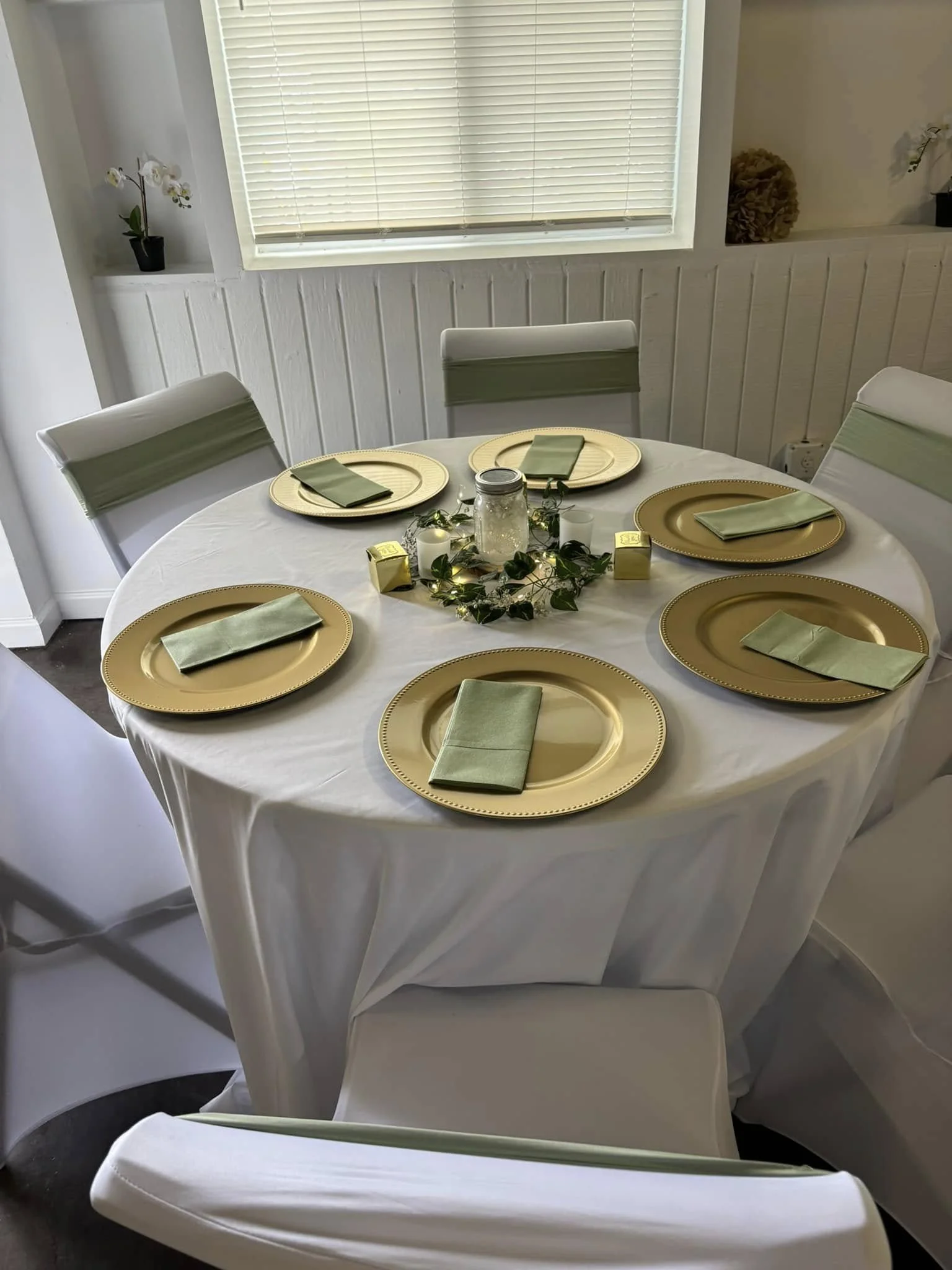 A round dining table set for six with gold chargers, green napkins, a glass jar centerpiece, and white candles. The table is covered with a white tablecloth and surrounded by white chairs with light green accents. A window with blinds is behind the t