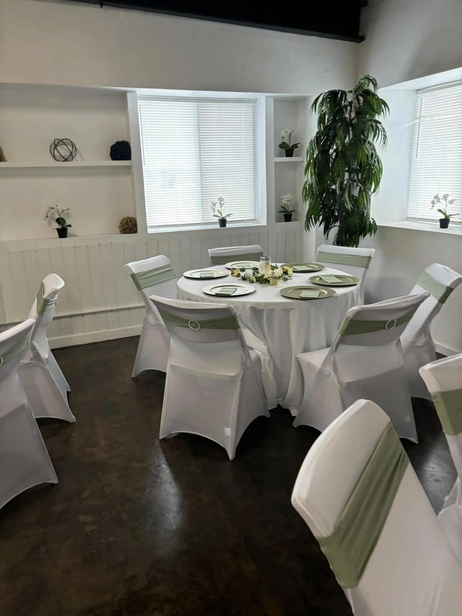 Round table set with white tablecloth, green napkins, and decorative centerpiece, surrounded by white chairs with green accents, in a decorated room with white walls, windows with blinds, and potted plants.