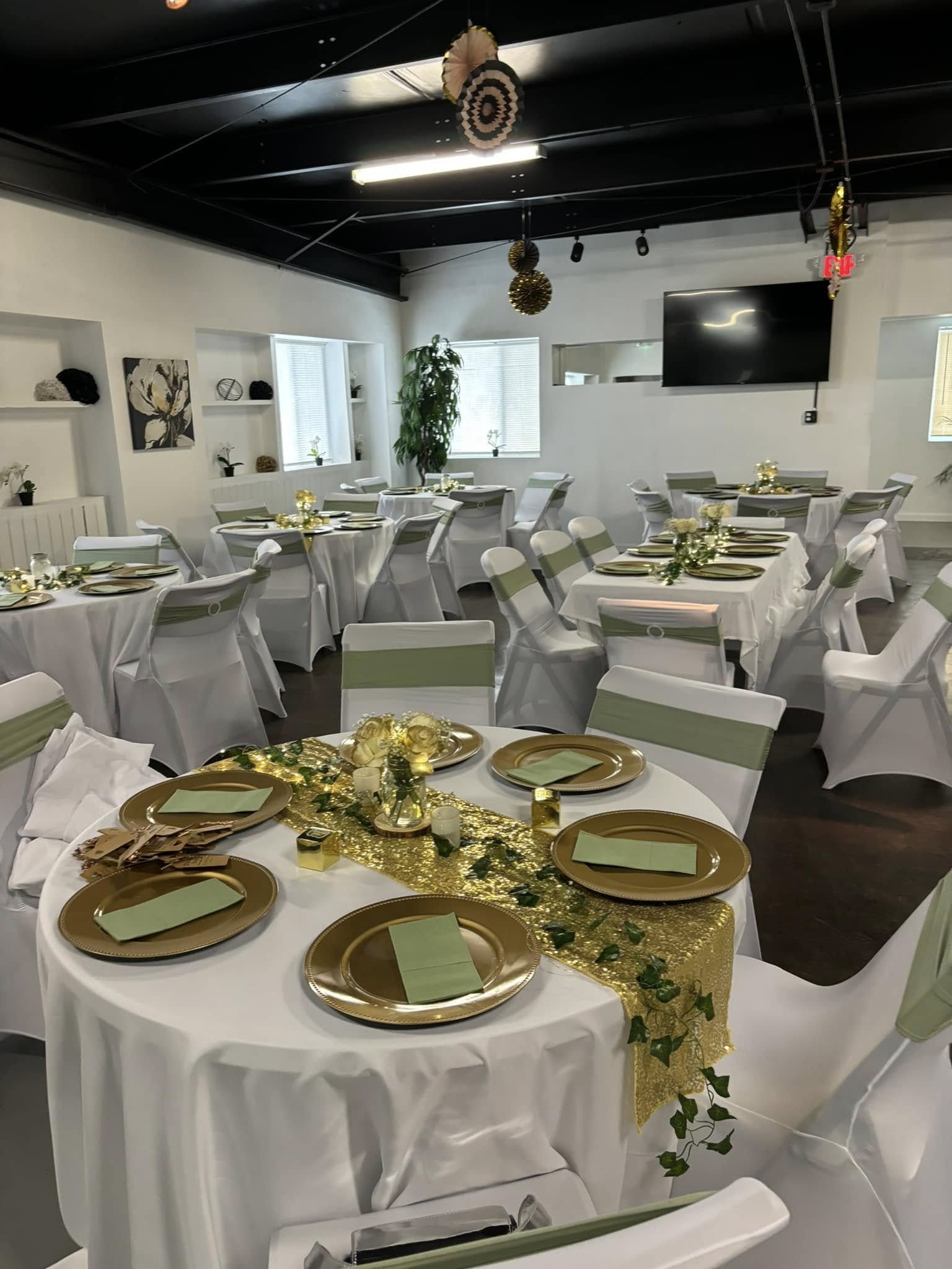 Event space decorated with white tablecloths, gold chargers, and green accents, featuring round tables with chairs, centerpieces, and hanging decorations.