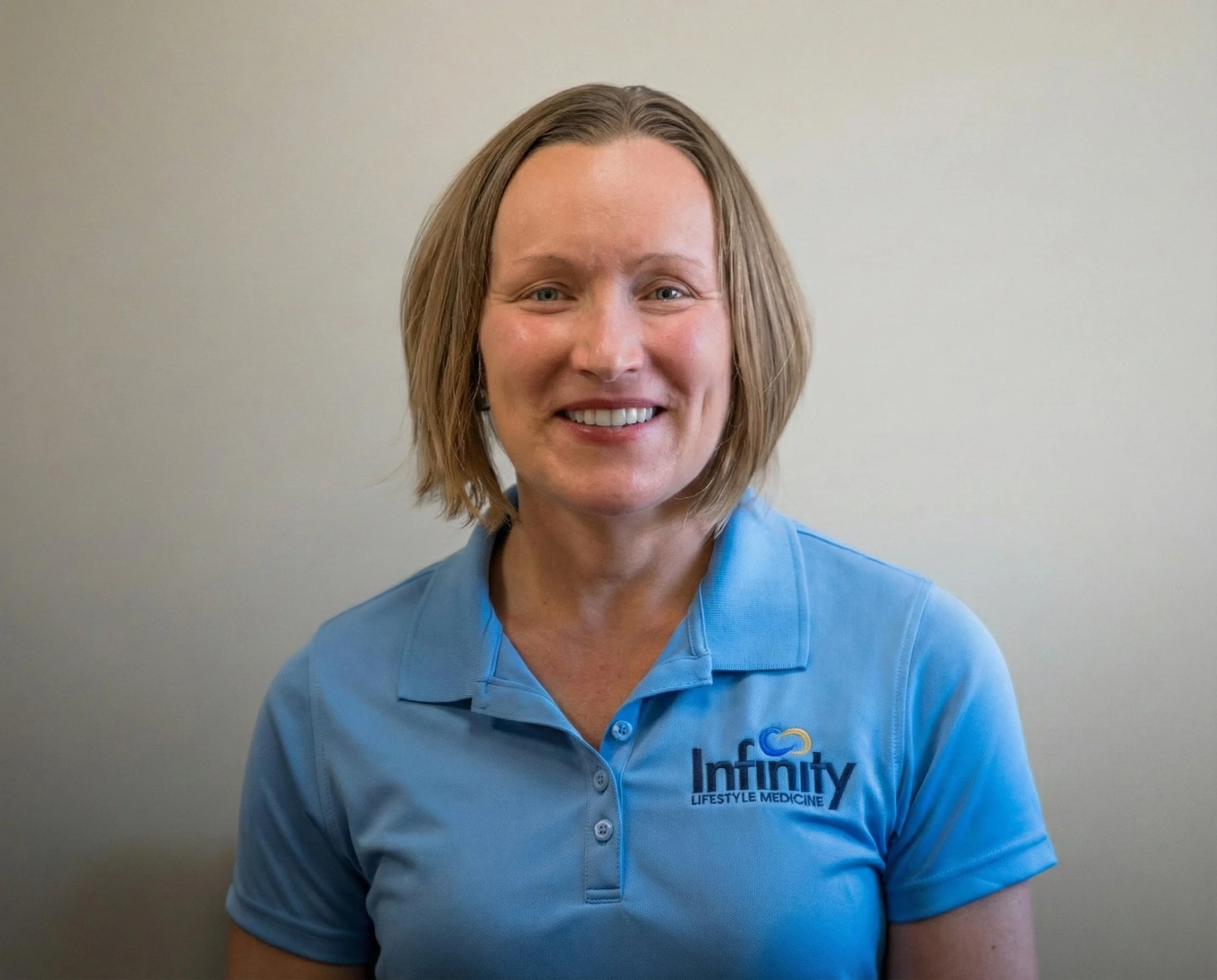 A woman with short brown hair and blue eyes, wearing a floral blouse and a gold necklace, smiling in front of a plain gray background.