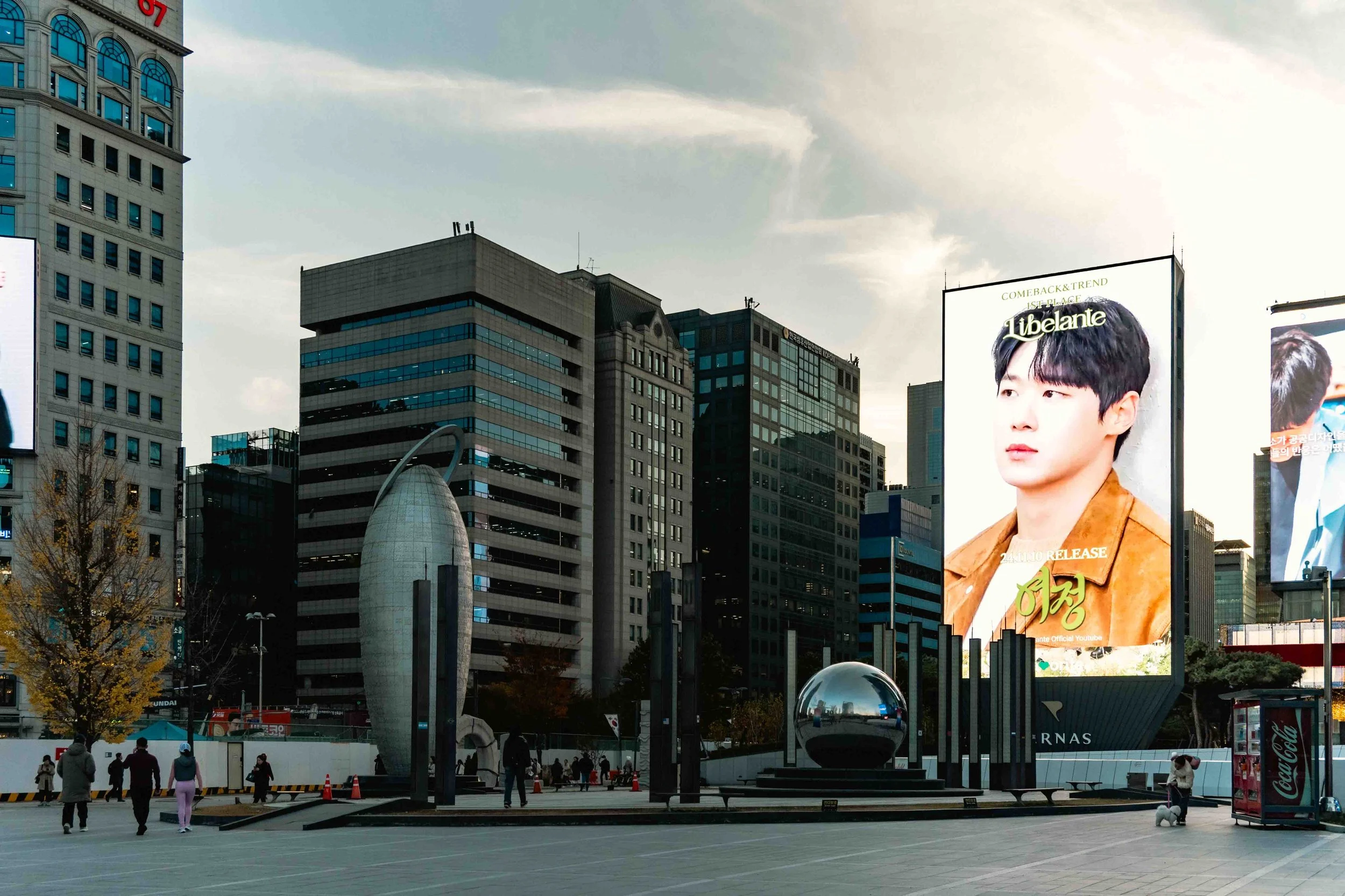 Public square in Gangnam with two billboards of Korean mal models with a few grey office buildings and metal sculptures.
