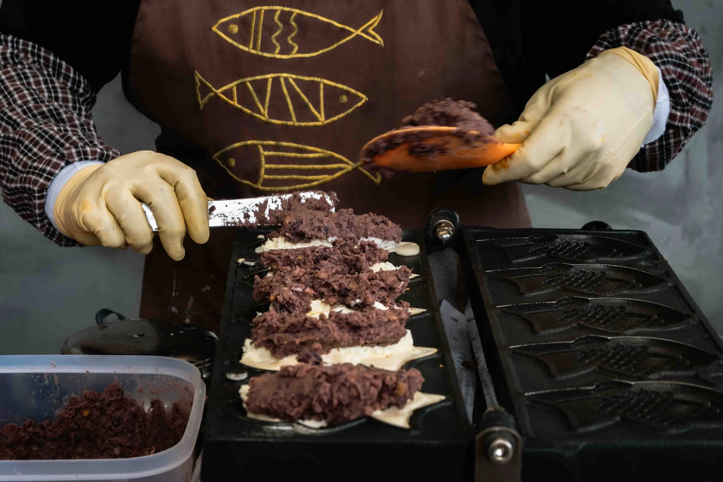 A person preparing Bungeoppang - a Korean fish-shaped pastry - with both of his hands to fill the pastry with red bean paste in a black skillet.