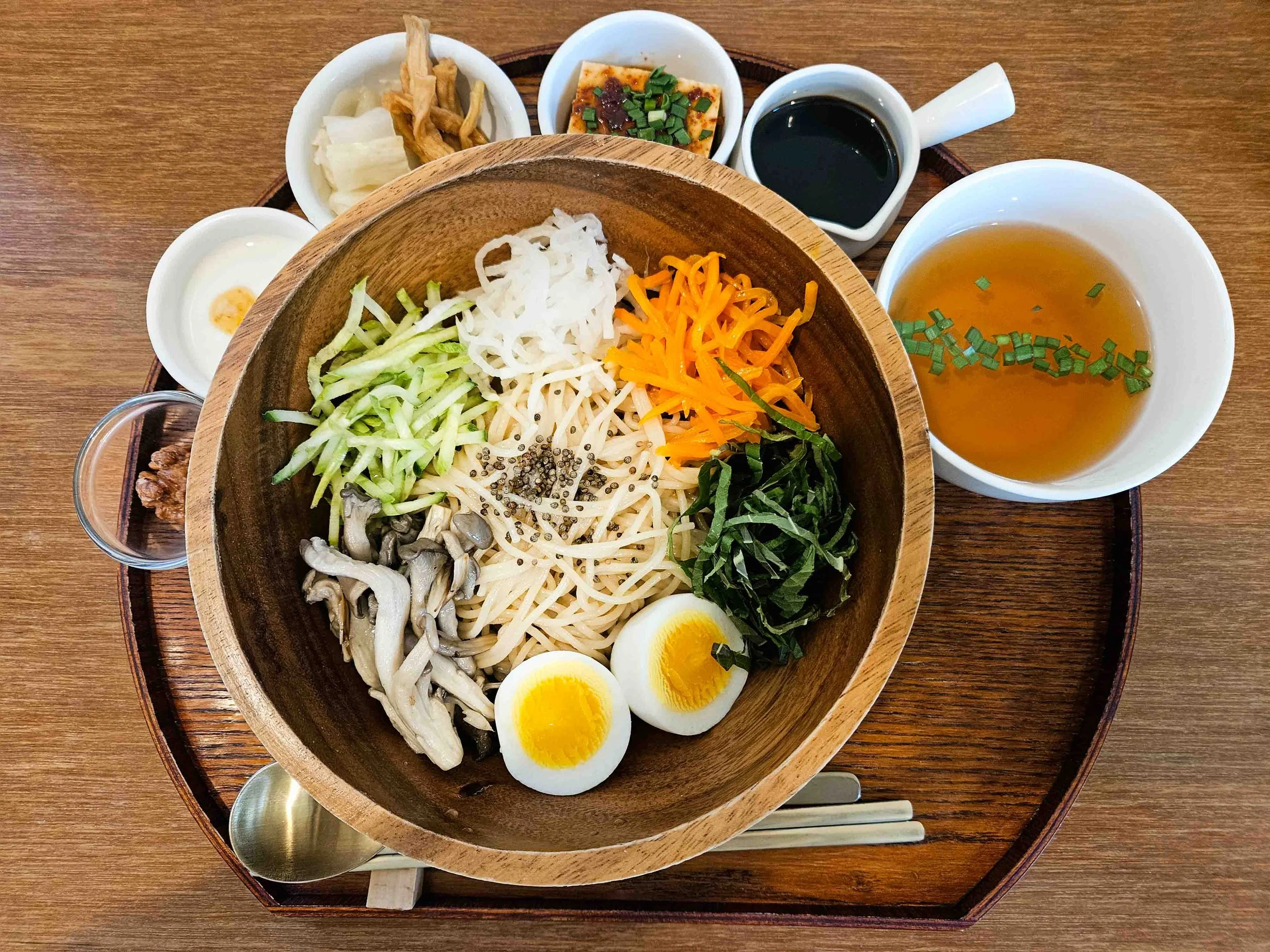 Bibimbap being served in a wooden bowl consisting of multiple colorful vegetables and side dishes in ceramic cups like tofu, walnuts, broth and soy sauce.