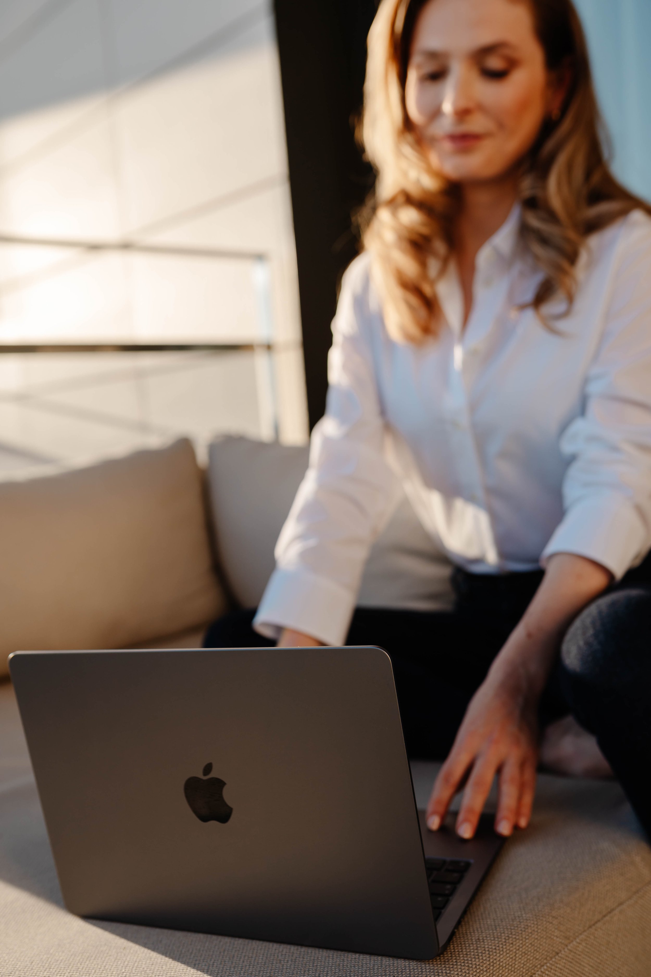 A woman in a white shirt sitting on a sofa with a laptop in front of her.