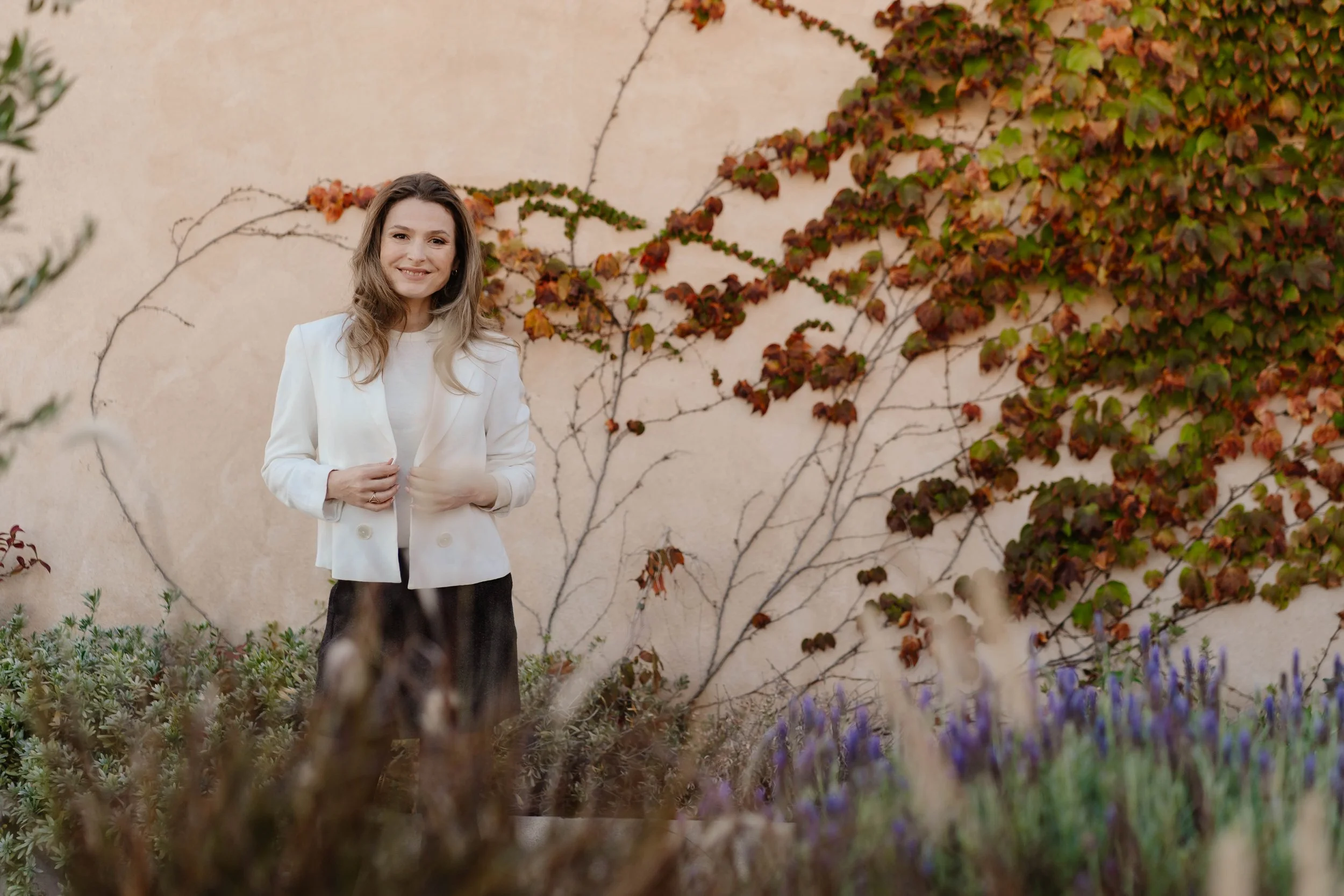 Eva Gaaff, website designer for coaches and creatives, wearing a white blazer and black skirt, standing outdoors in front of a beige wall with climbing red and green ivy, smiling at the camera.