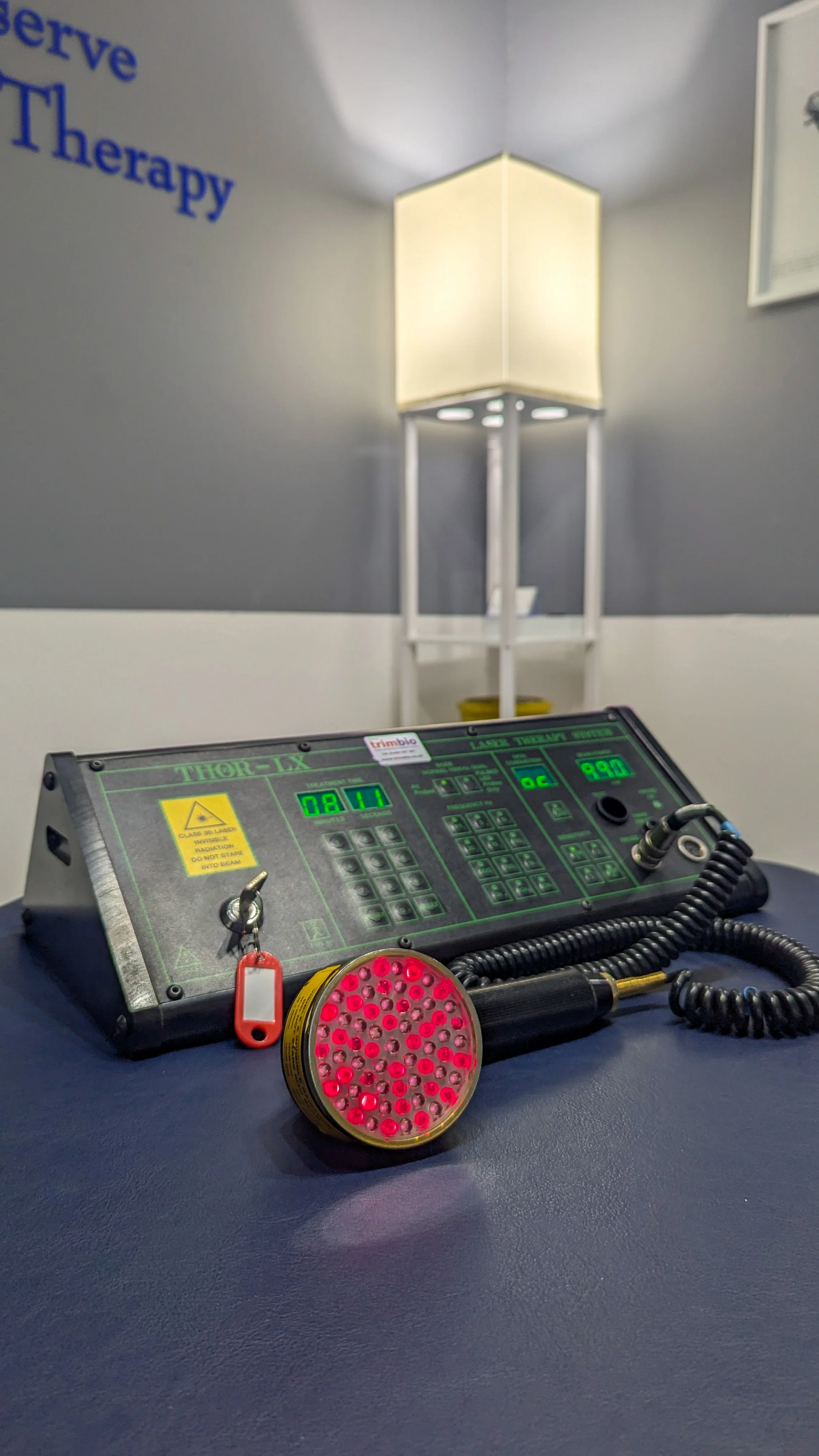 Laser therapy device on a table in a medical office, with a lighted lamp and framed certificate in the background.