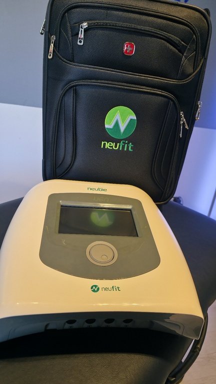 A black bag with a red logo, standing behind a medical device branded 'neufit', which has a large display screen and a circular control button, on a black surface.