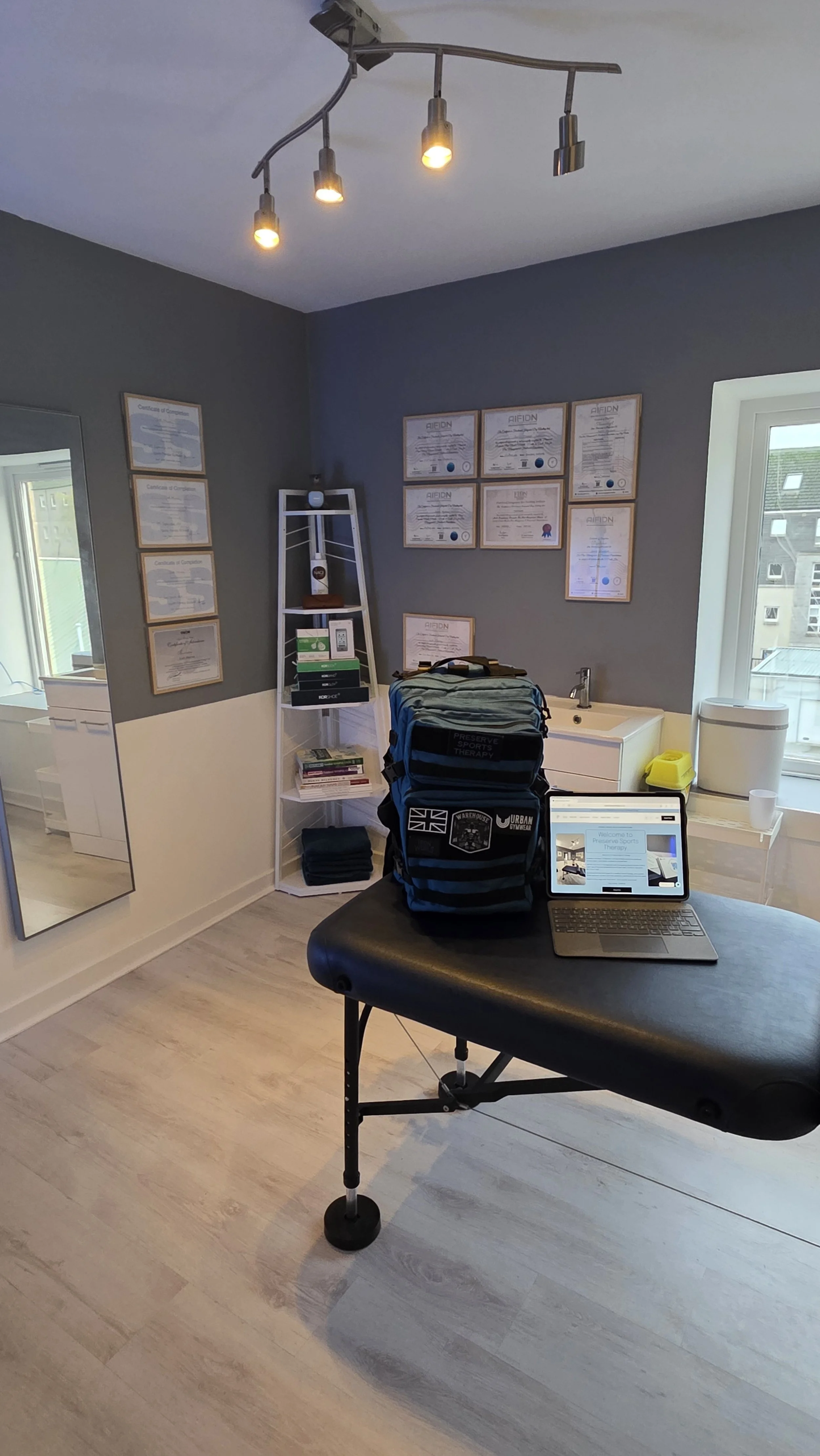 A room with a massage or treatment table, a laptop, a backpack, and framed certificates on the wall.