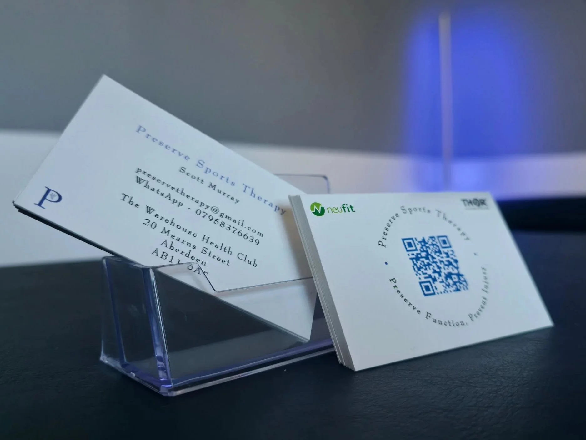 Business cards for Preserve Sports Therapy featuring a QR code, contact information, and branding from Neufit, displayed on a black surface with a soft blue light in the background.