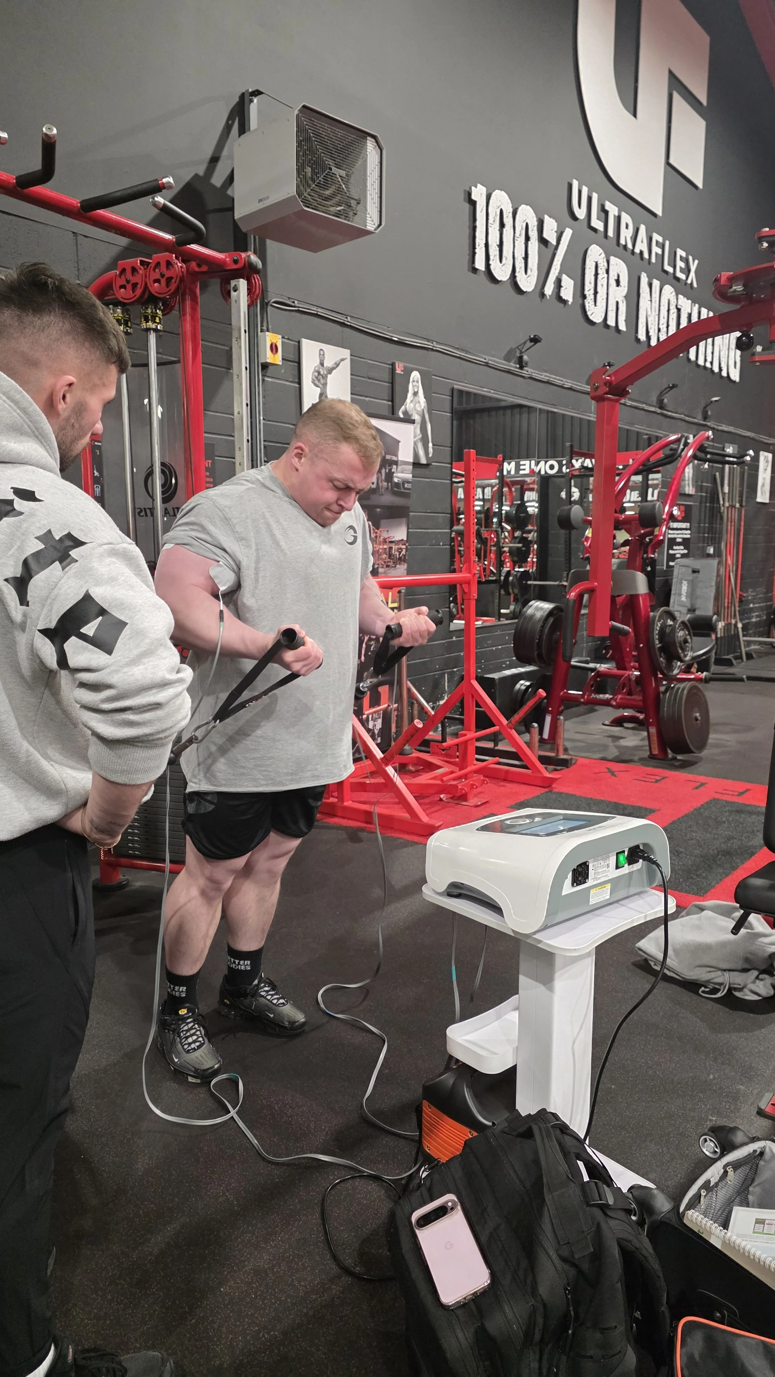 Two men in a gym using electrical stimulation equipment on their arms, with gym equipment and weights in the background. Khifie West, Chris McCreadie, Neubie, Biceps