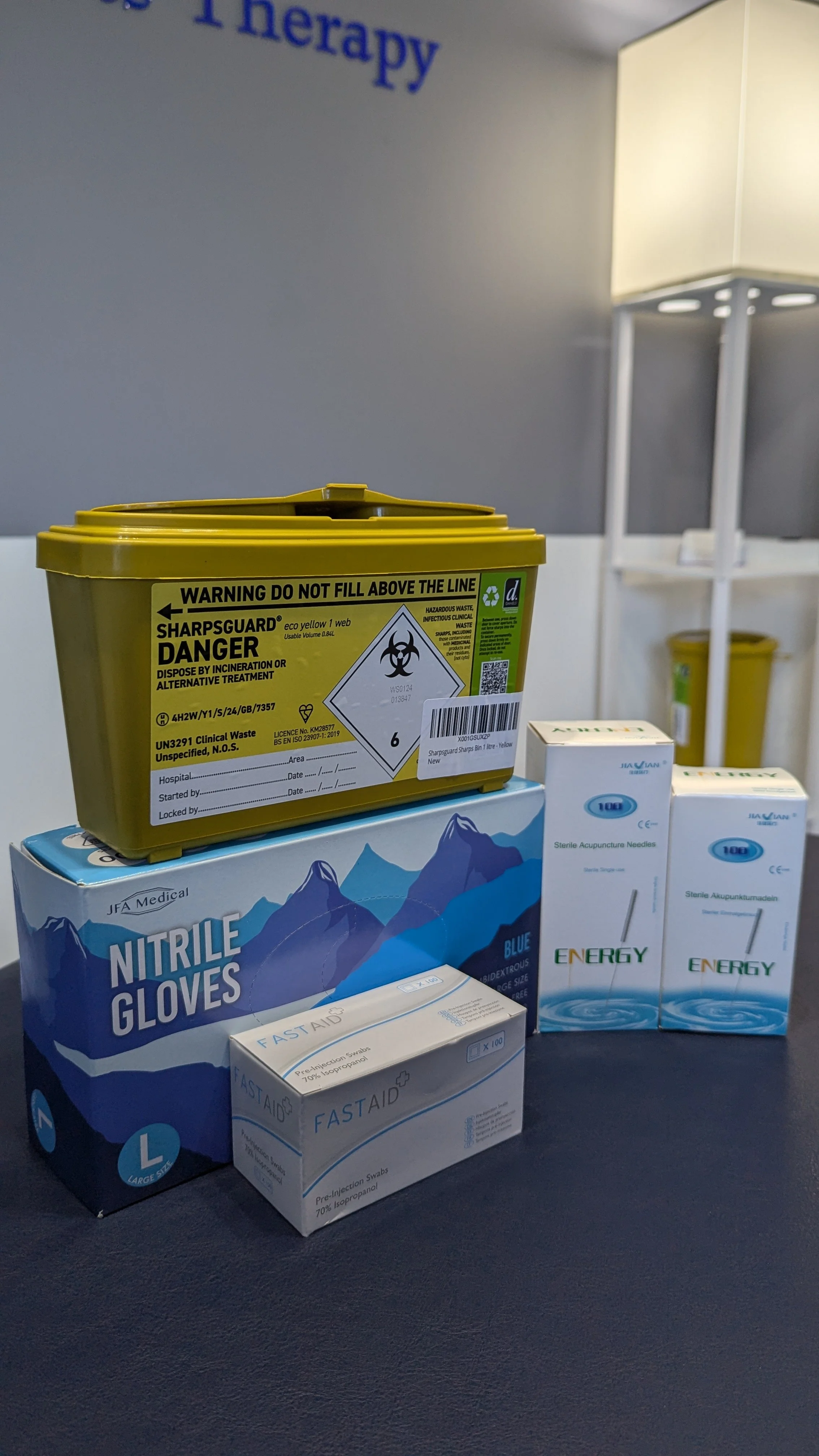 Medical supplies including a yellow biohazard sharps container, blue nitrile gloves, boxes of acupuncture needles, and boxes of pre-injection swabs on a dark surface.