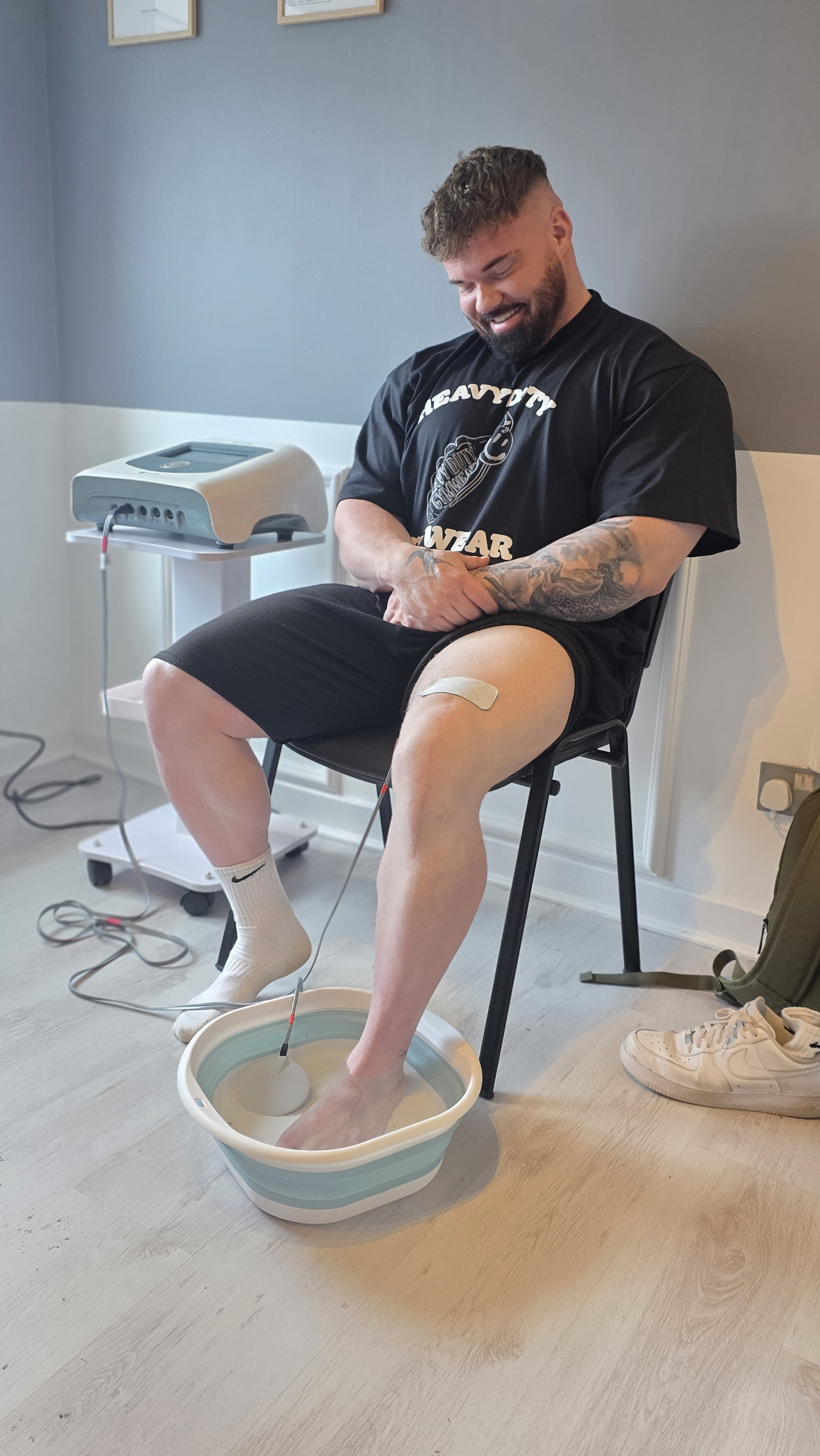 A man sitting on a black chair with his right leg in a white basin filled with water, receiving an electrotherapy treatment. He is smiling, has a tattooed arm, and a bandage on his thigh. There is a medical device beside him connected with wires to his leg. The room has light-colored walls, some framed pictures, and medical equipment.