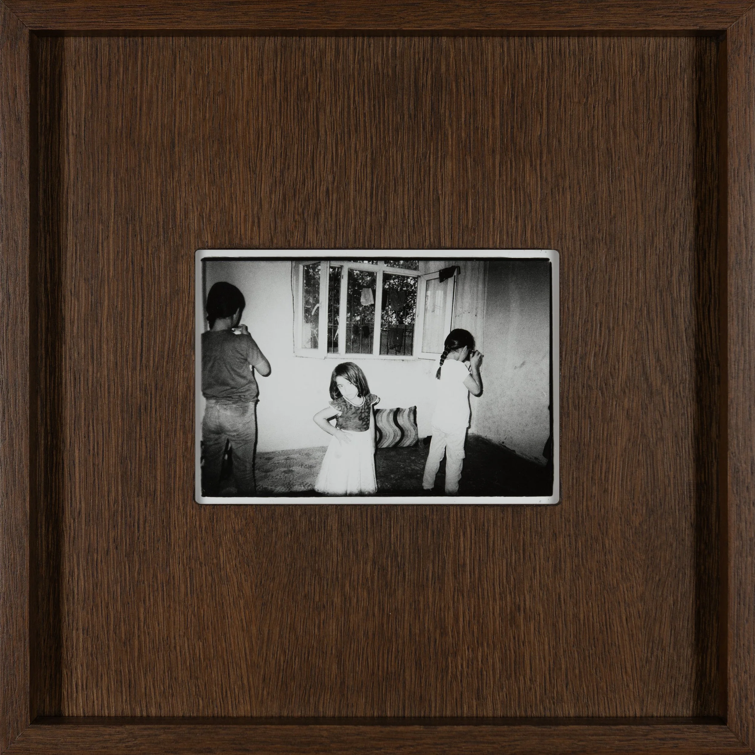 A 13-year-old child stands in the foreground of a dim, modest room, captured in black and white.