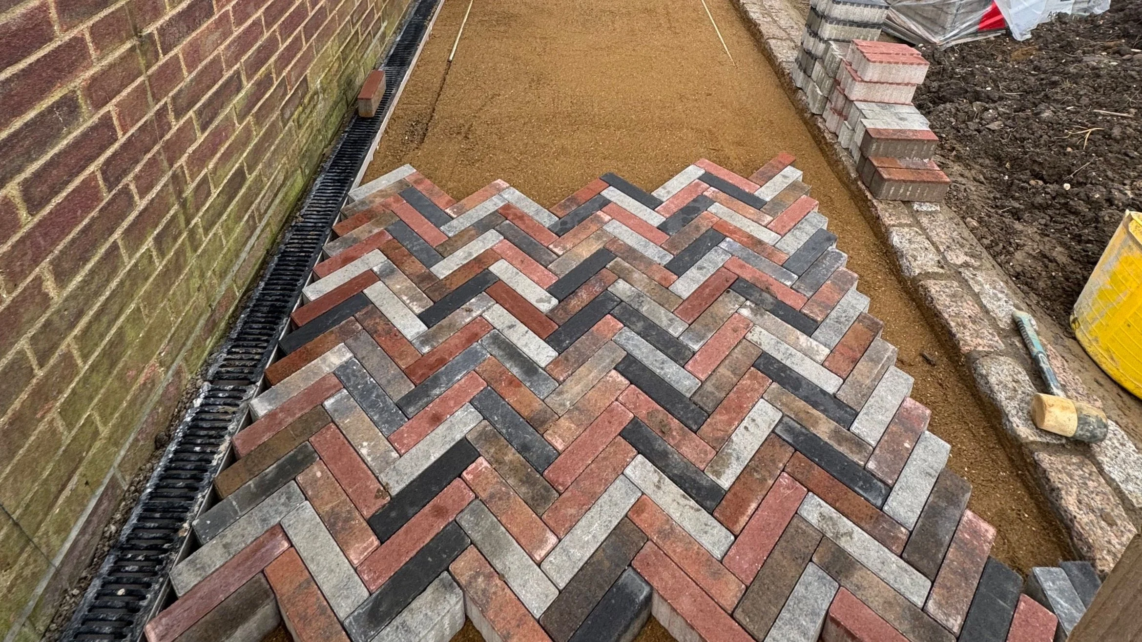 Hard landscaping services in progress showing a herringbone pattern block paving installation beside a brick wall, with drainage channels and edging in place.