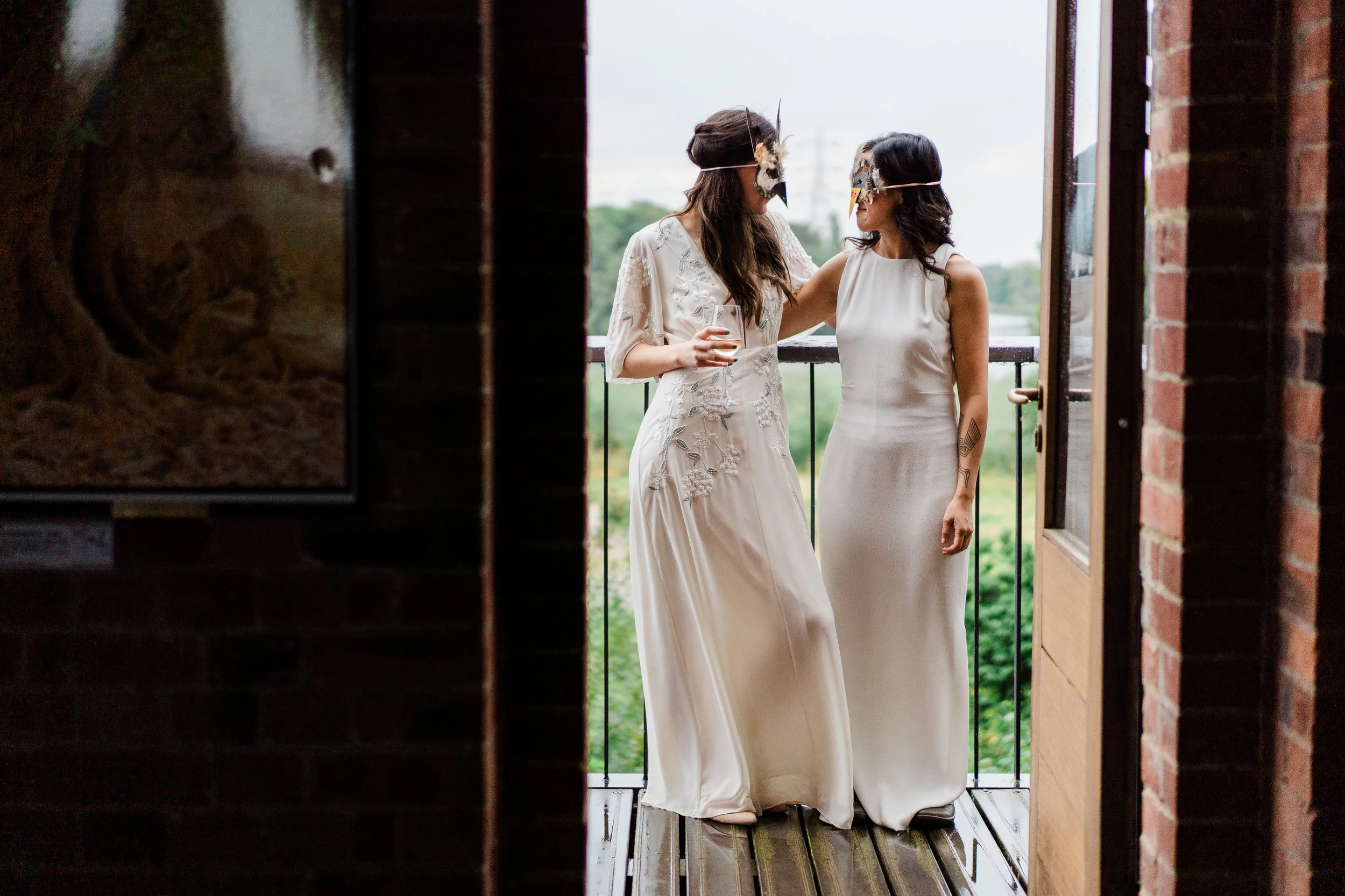 Two women wearing wedding dresses with masks, standing on a balcony, talking and holding a glass of wine.