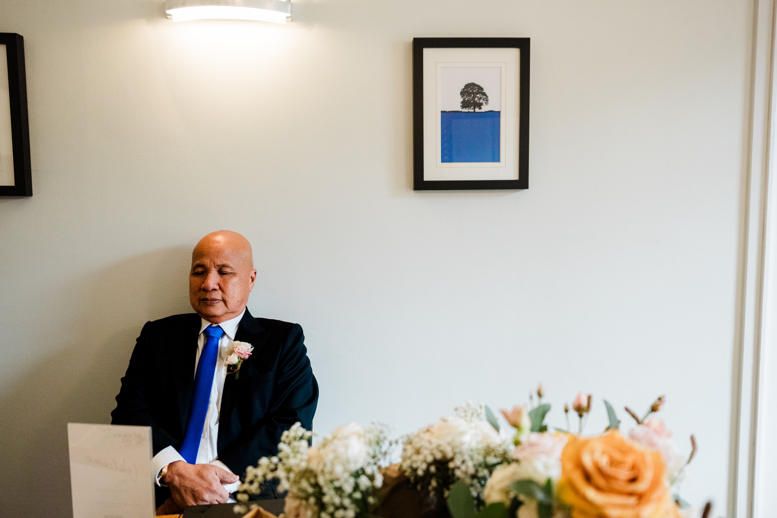 An older man in a black suit with a blue tie and pink boutonniere sitting with eyes closed in a room decorated with flowers, on wall is a framed picture of a tree and sky.
