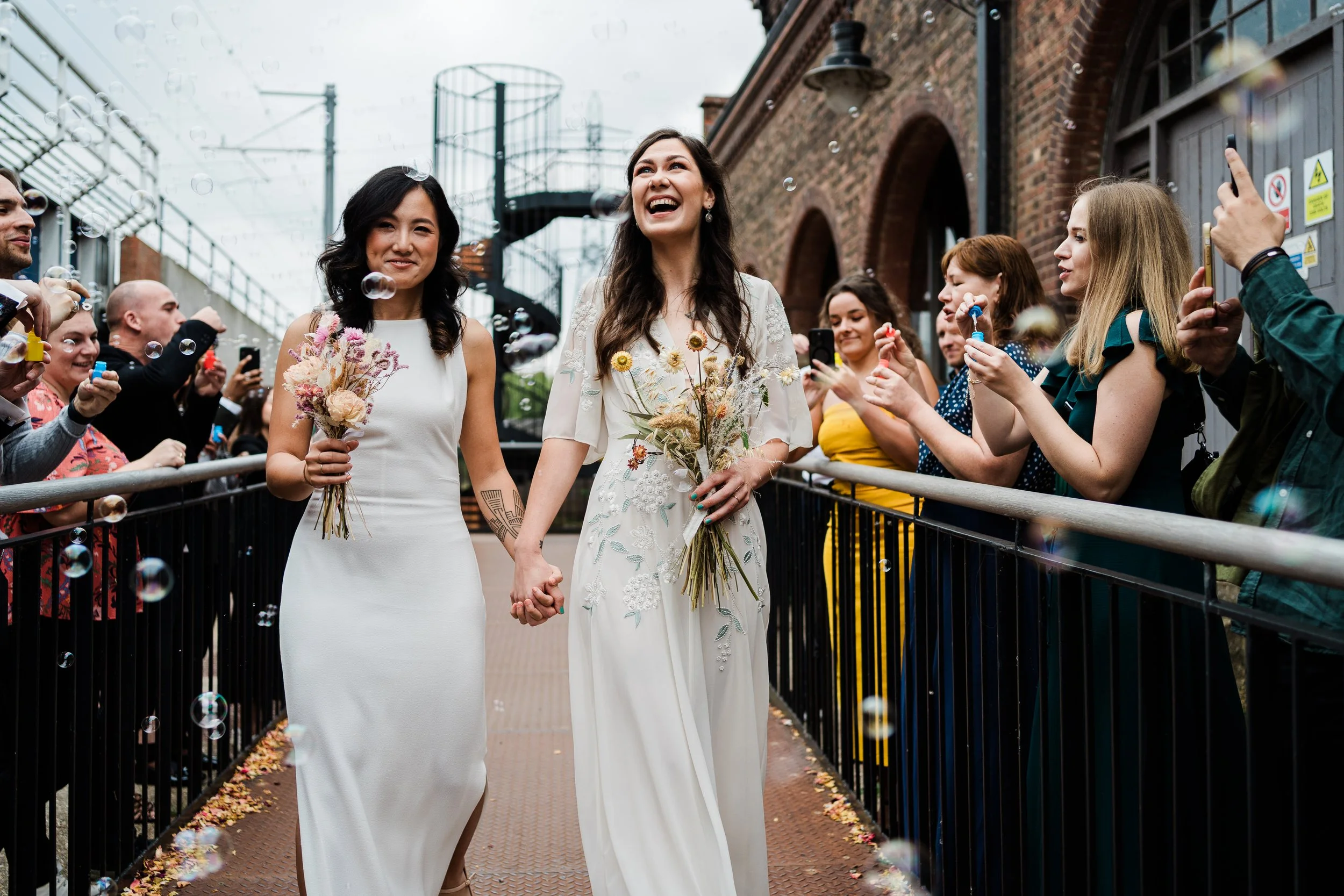 Two women in wedding dresses holding hands and bouquets, smiling and walking through a crowd of friends and family celebrating with bubbles outside near a brick building.