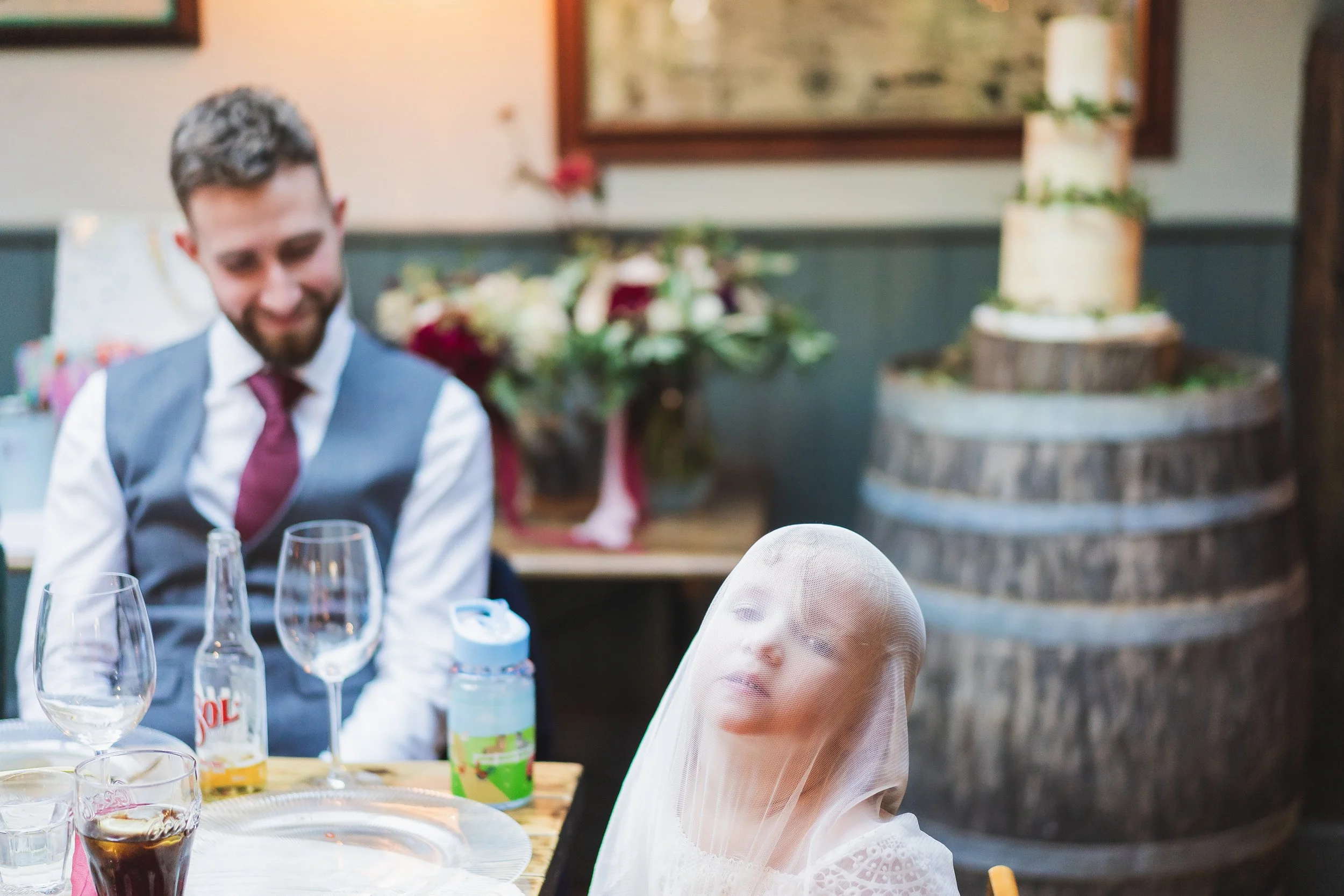A young girl with a veil over her head sitting at a table with empty glasses and bottled drinks, looking to the side. Behind her and to the left is a man in a suit and tie smiling, with a decorated wedding cake visible in the background.